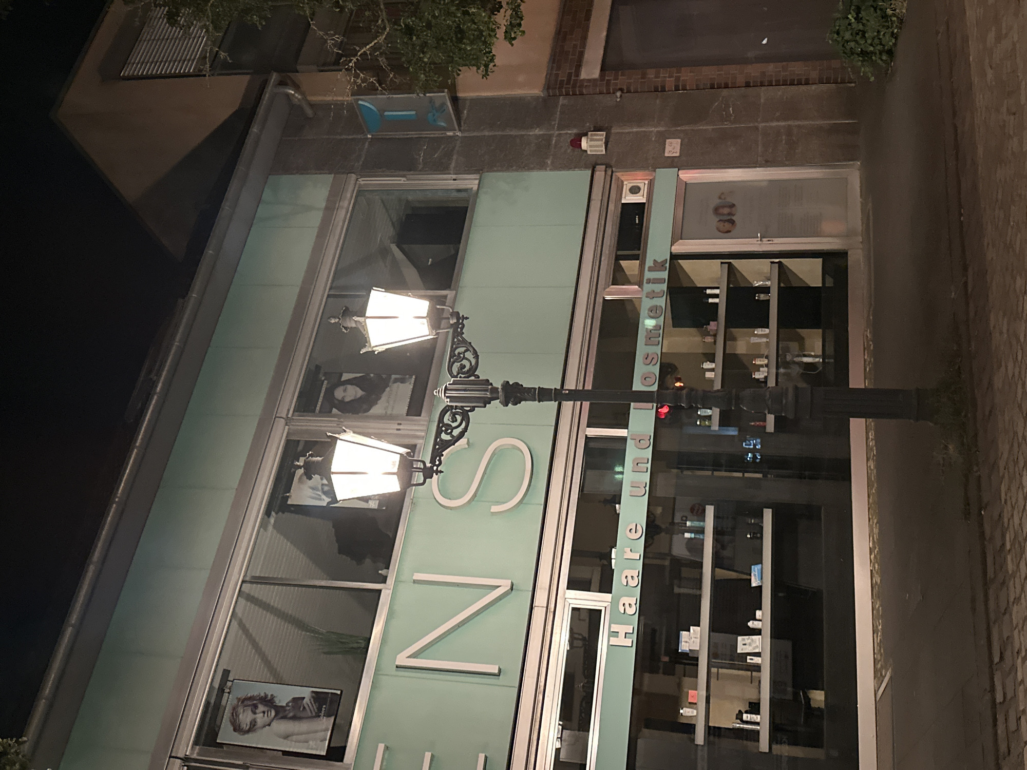 The image depicts the exterior of a store at night in Krefeld, Germany. The storefront features a light green facade with large windows. Above the windows, prominent white lettering reads "ENS". A smaller sign below the windows says "Haare und Kosmetik", indicating the business is a hair and cosmetics salon. The window displays shelves stocked with various bottles and containers, presumably beauty products. A street lamp with two illuminated lights stands in front of the building. The street in front of the store is paved with cobblestones. The building to the right of the store is made of brick. The overall atmosphere suggests an urban commercial setting illuminated by artificial light.