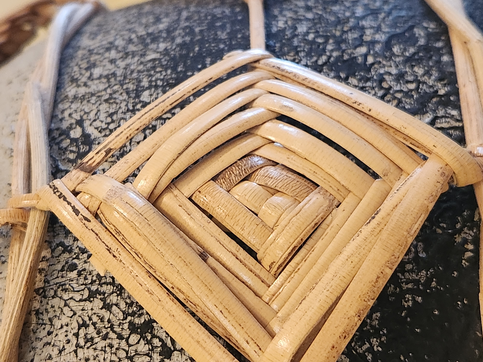 The image is a close-up, angled shot of a woven wicker or rattan detail on a dark, textured object, possibly a pot or vase. The weaving is in a diamond pattern, with the strands of the material layered and interwoven to create a recessed square shape at the center. The wicker is a light, natural wood color and shows some natural variations in tone and texture. The dark object beneath the weaving has a speckled, somewhat rough surface. The scene appears to be an indoor or outdoor setting, though no specific location in Maitland, United States of America can be identified from the visual information alone.