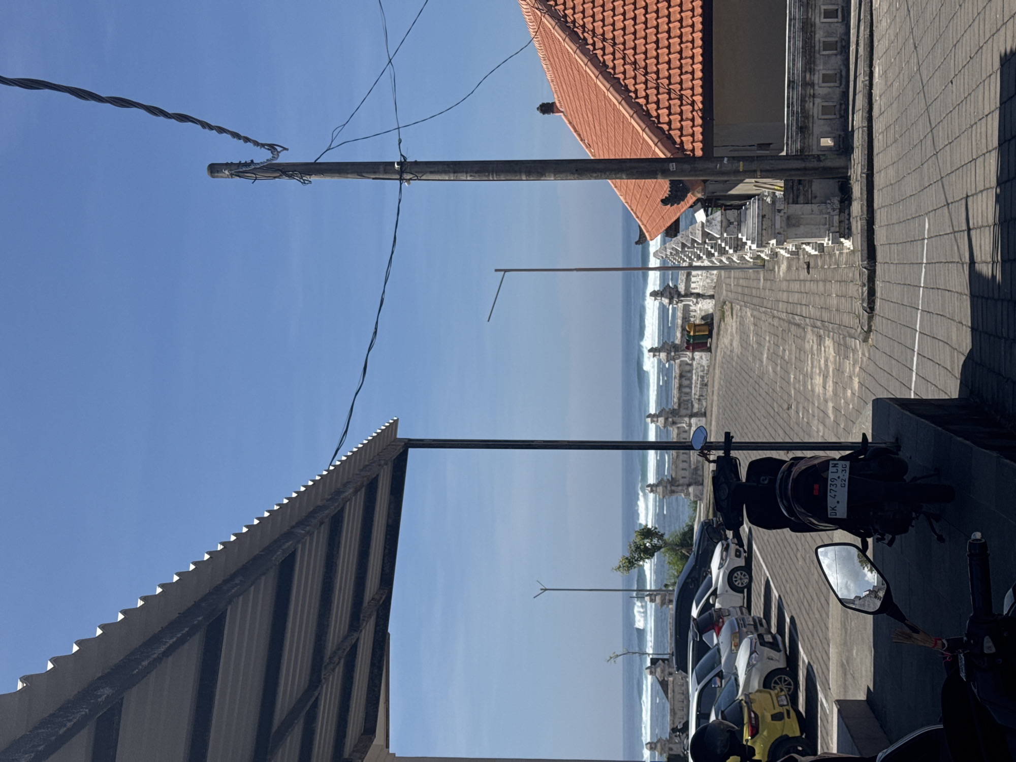 A street scene in an unknown city, unknown, shows a view towards the ocean on a clear day. Utility poles and wires are prominent. A tiled roof is partially visible at the upper right, and a corrugated metal overhang is visible at the lower left. Parked cars and motorcycles are visible along the right side of the street. A tiled sidewalk and low ornamental wall line the edge of the street. Ocean waves are visible in the background.