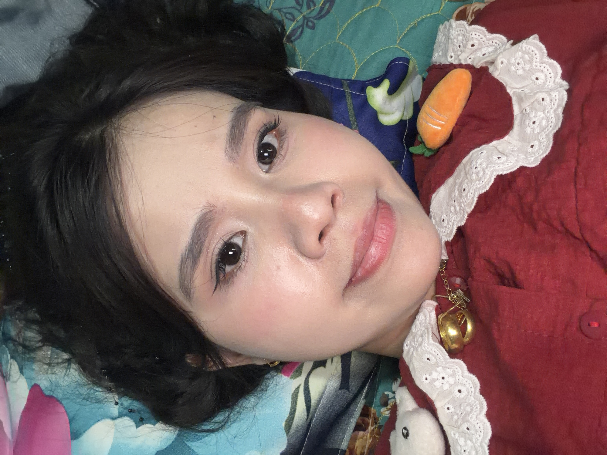 A person is lying on a patterned bedspread, with their head resting on a blue pillow featuring green floral patterns. The person has dark hair, light skin, dark eyes, and defined eyebrows and eyelashes. They are wearing a red garment trimmed with white lace. A small, plush carrot is positioned near the neckline of the garment, and a gold bell earring hangs from the person's ear. A small, plush white bunny is also visible near the garment. The location is not determinable from the image.