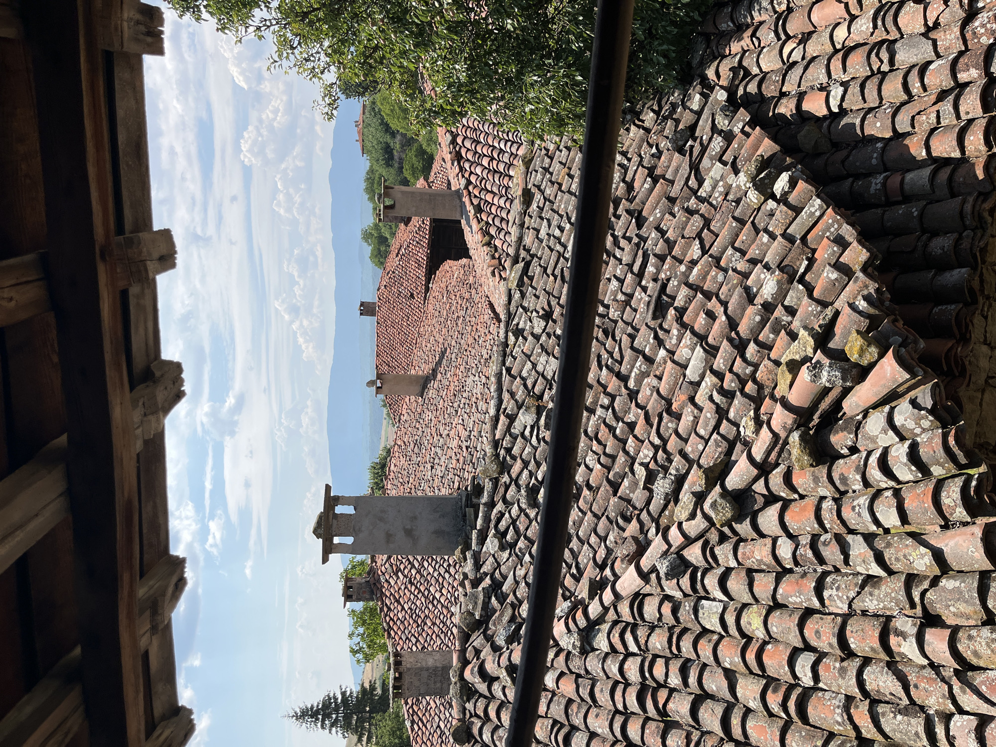 The image, taken from a high vantage point within a wooden structure, showcases a panoramic view of Zheravna, Bulgaria. The foreground is dominated by aged, red-tiled rooftops, some of which are partially obscured by a dark horizontal beam. Several stone chimneys, varying in height and design, punctuate the tiled landscape. In the mid-ground, more red-tiled roofs and glimpses of building facades are visible amidst lush green trees. The background reveals rolling hills and distant mountains under a bright blue sky dotted with white, fluffy clouds. The overall scene depicts a traditional, historic Balkan village bathed in natural light.