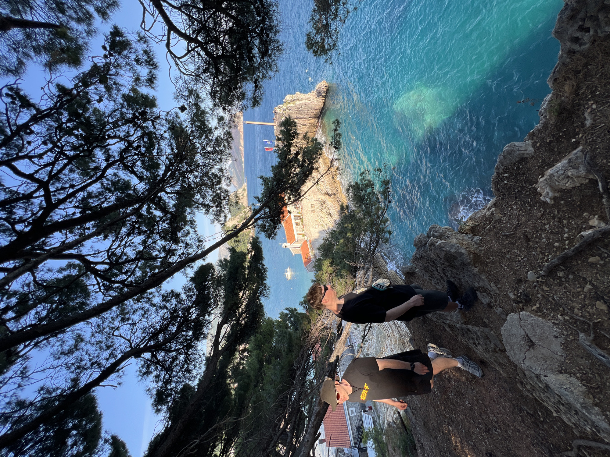 Two people stand on a rocky cliff edge overlooking the Adriatic Sea in Petrovac Centar, Montenegro. The water is turquoise and clear, revealing underwater features. A small island with a stone building and a dock featuring two red flags extends into the sea. Trees frame the scene, obscuring parts of the sky. The person closest to the camera wears a dark shirt, khaki hat, red sunglasses, and dark shorts and grey sneakers. The second person is wearing a black shirt, dark pants, dark sunglasses, and black shoes. Buildings with red roofs are visible in the background.