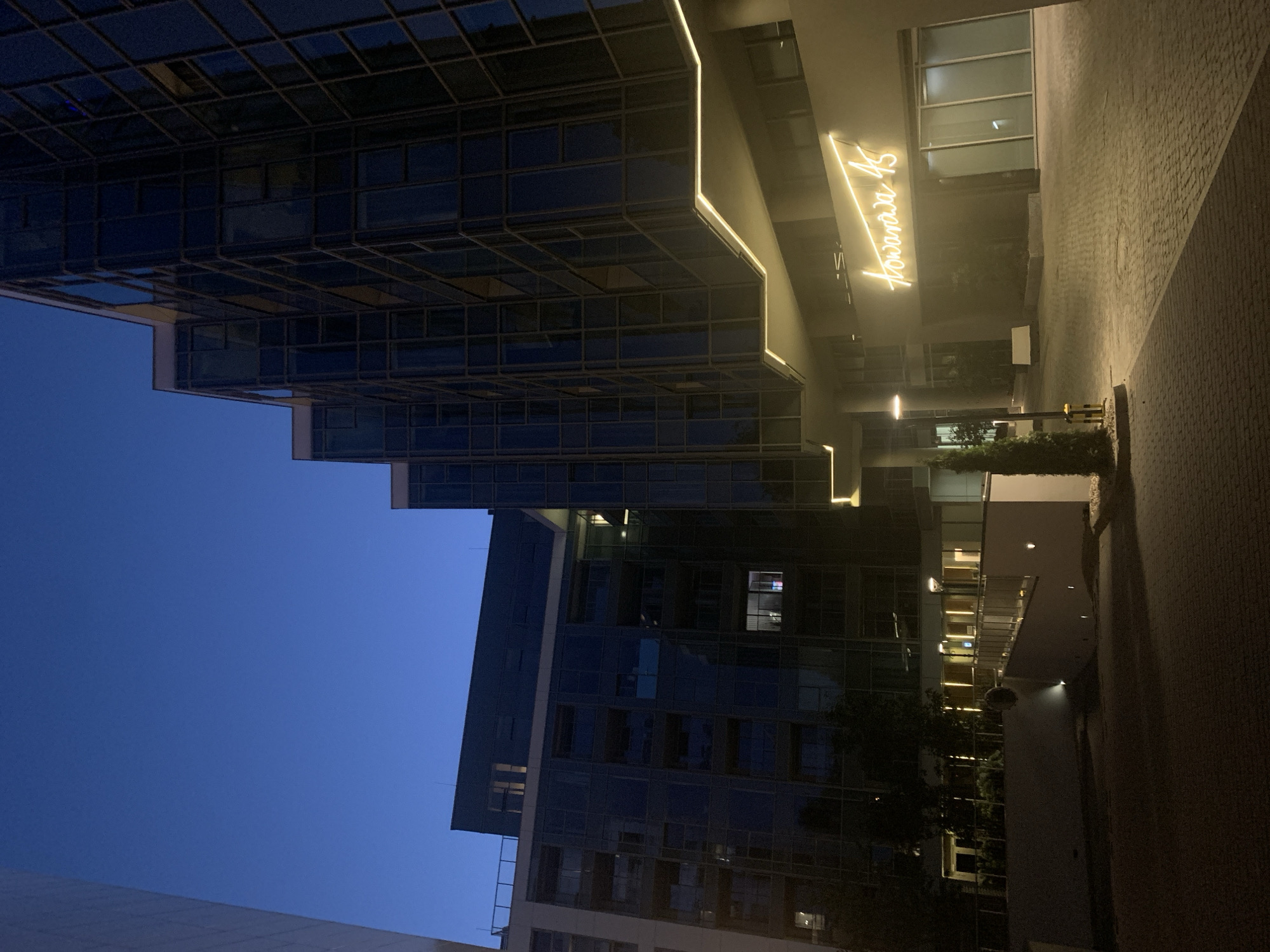 This is a night exterior photograph of the Aura Apartments building located at Tęczowa 45 in Poznan, Poland. The building features a modern architectural design with large glass windows and balconies. The sky is a deep blue, indicating twilight or early night. Subtle yellow lighting is visible along the building's facade and at the entrance, illuminating the signage which reads "Tęczowa 45". The ground in front of the building is paved with cobblestones. A lamppost stands to the left of the entrance, and a small evergreen tree is planted nearby. There are no people visible in the image. The overall impression is one of a contemporary urban setting at dusk.