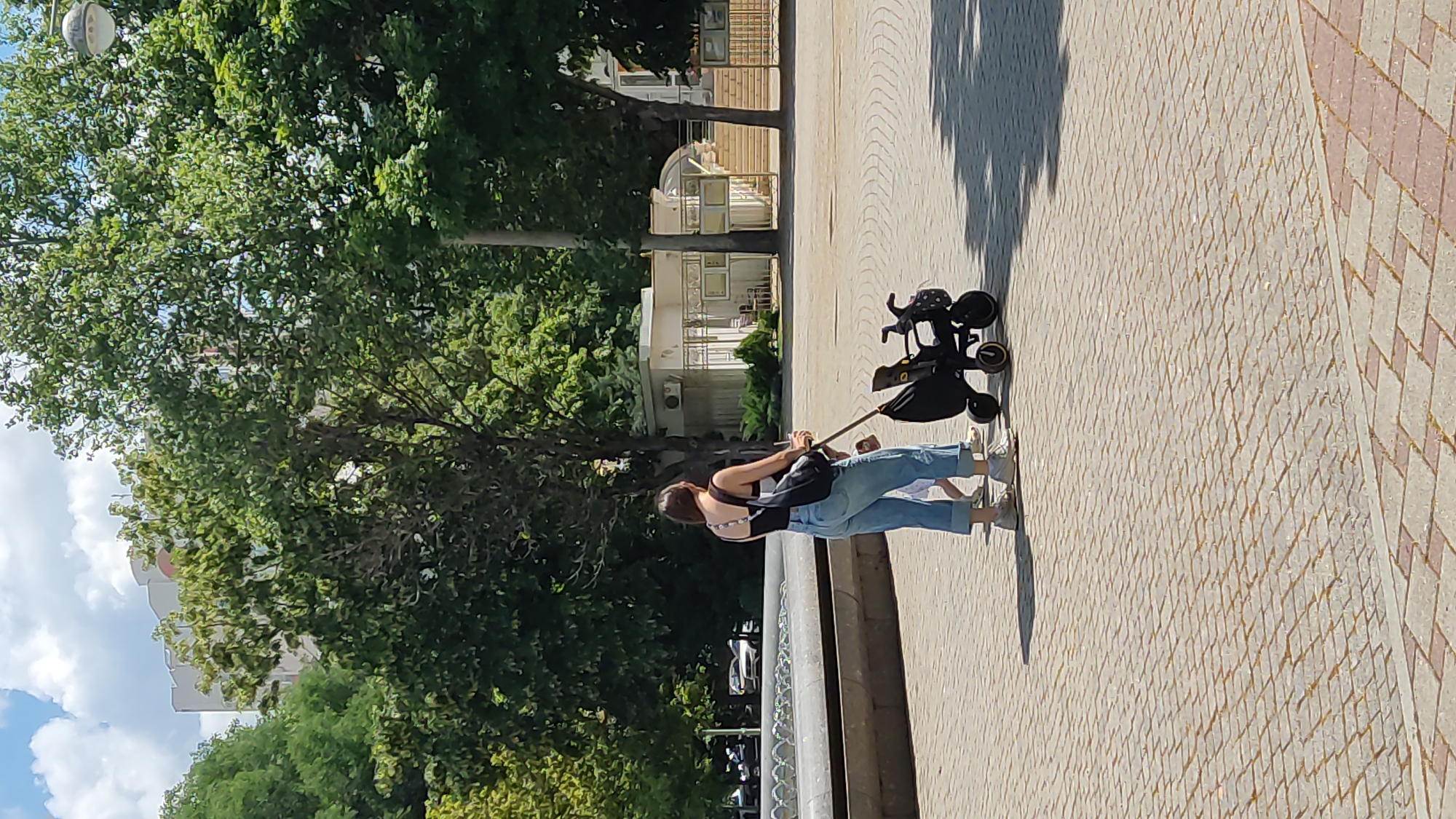 The image shows a person from behind, pushing a black stroller on a paved walkway. The person is wearing a black top with a patterned strap and light blue jeans. They are also wearing white sneakers. The stroller has a black frame and several wheels. The walkway is made of light-colored rectangular pavers arranged in a diamond pattern, with some darker, reddish pavers interspersed. A shadow of the person and stroller is cast on the walkway, indicating it is daytime.
In the background, there are green trees with lush foliage, a white building with multiple windows, and a section of a building with a curved, glass-enclosed entrance. The sky is visible above the trees and building, with some white clouds. The overall scene appears to be an outdoor public space, possibly a park or promenade.
The image is taken in Krasnodar, Russia. The weather appears to be sunny, given the shadows and the bright lighting. The person's posture suggests they are casually walking and pushing the stroller. There is no visible text in the image.