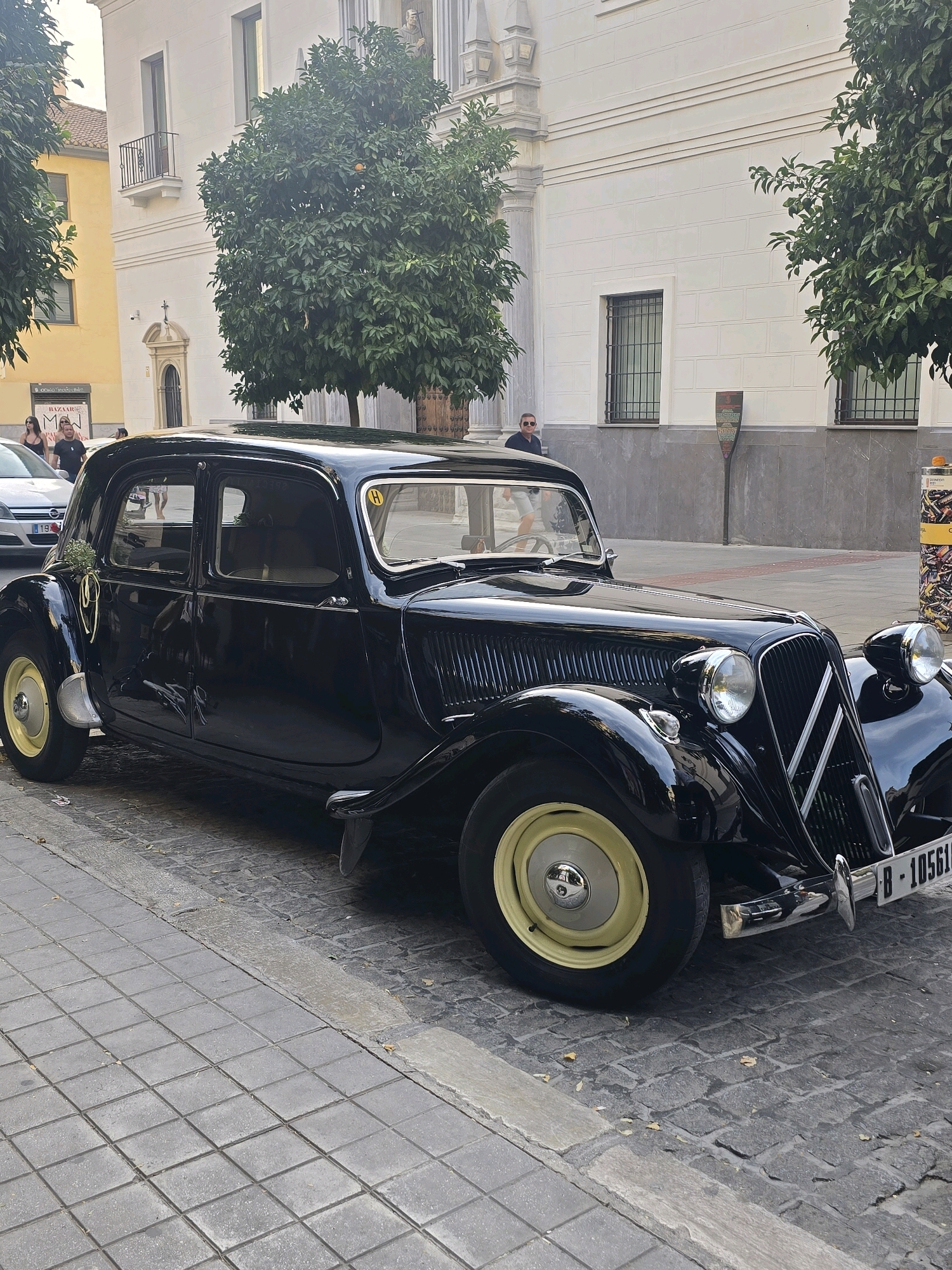 The image shows a vintage black car parked on a cobblestone street in Granada, Spain. The car, a Citroën Traction Avant, is ornate with chrome accents and pale yellow hubcaps. A small bouquet of white flowers tied with yellow ribbon is attached to the rear fender. In the background, a white building with a classical architectural style stands, featuring balconies and arched windows. Orange trees with some fruit are visible to the left and right of the car, lining the street. People are seen in the background, some walking and others standing near the building. A man in a dark shirt and sunglasses is standing near the car, looking towards the building. The scene appears to be a sunny day, with soft lighting illuminating the street and buildings.
