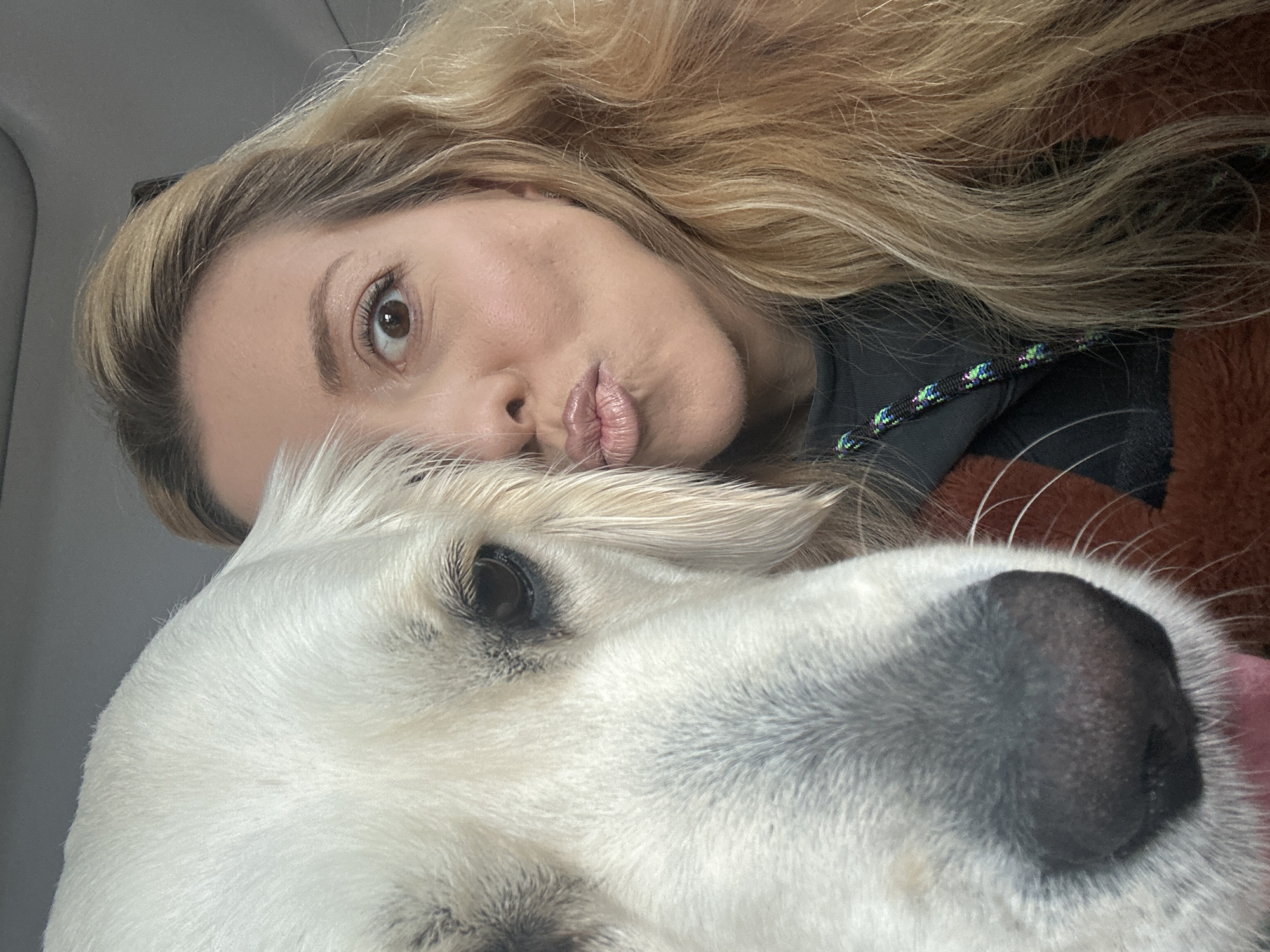 A selfie-style photograph shows a woman with long blonde hair and a white dog. The woman is partially visible at the top, her face angled towards the camera with pursed lips. The dog occupies the lower portion of the frame, with a large black nose and one eye visible. The background is indistinct, appearing to be the interior of a vehicle. The location is not identifiable as London, United Kingdom, based on the visual elements present in the image.