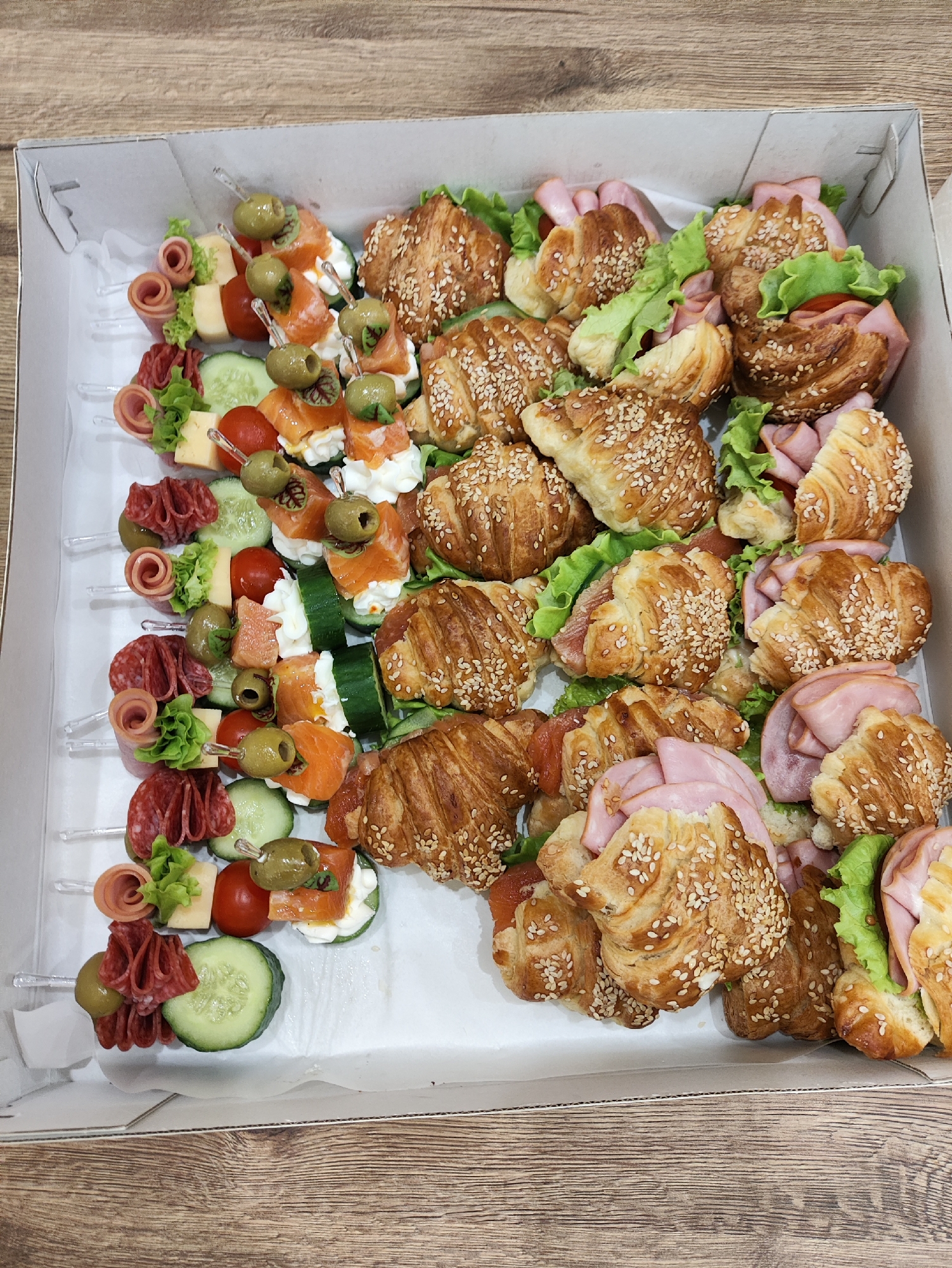 This is a top-down, eye-level shot of a catering box filled with various appetizers. The box contains rows of mini croissants filled with ham and lettuce, as well as skewers with salami, olives, cheese, cherry tomatoes, cucumber slices, and what appears to be smoked salmon with cream cheese. The croissants are golden brown and sprinkled with sesame seeds. The skewers are arranged on the left side of the box, with the croissants filling the right and bottom portions. The overall impression is a well-prepared assortment of finger foods, likely for a party or gathering. The setting appears to be indoors, with a wooden surface visible beneath the open box. There is no visible text in the image. The lighting suggests it could be daytime, with natural light perhaps supplemented by artificial sources. The scene is set in Старое Мартьяново, Russia.