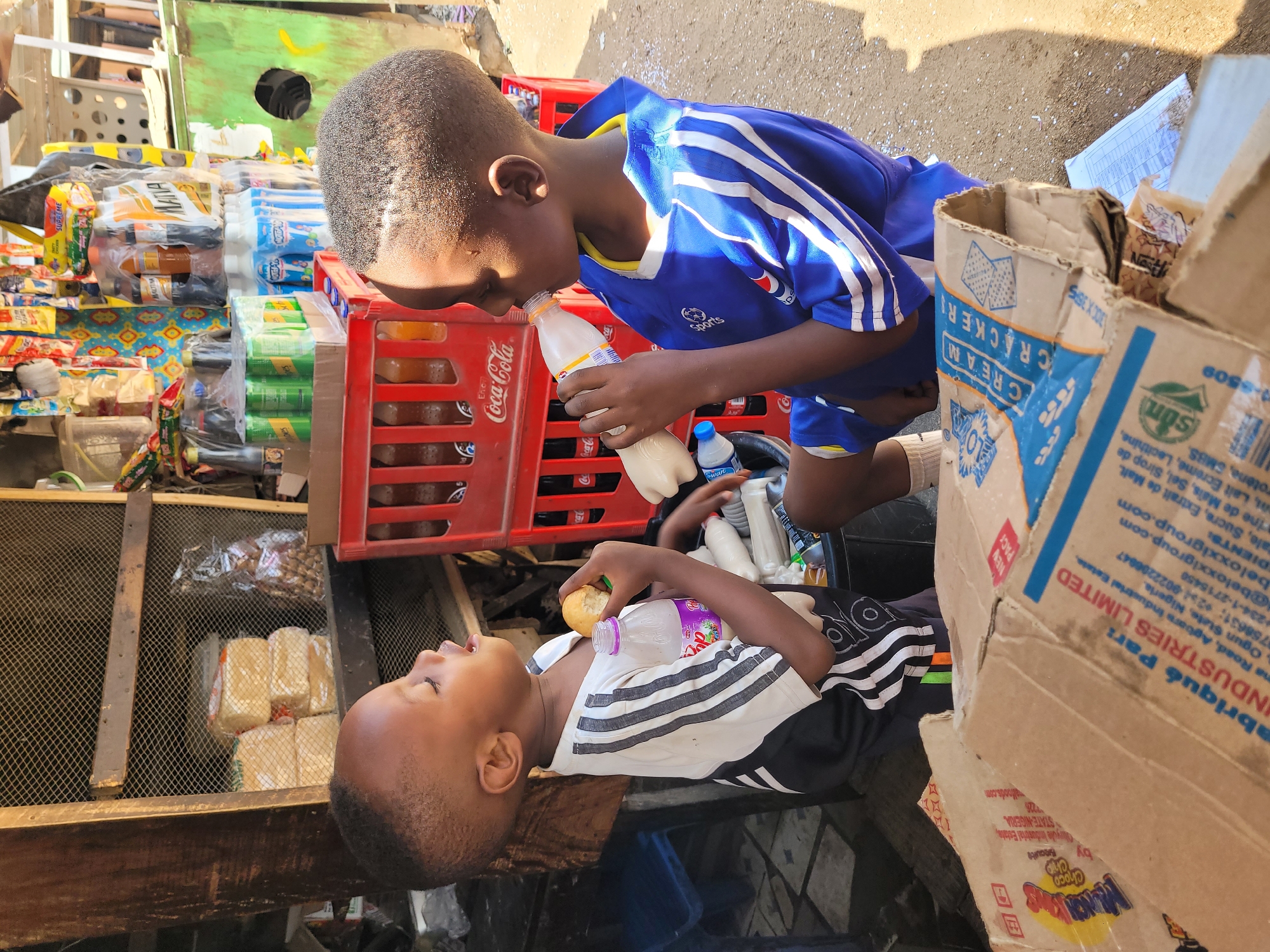 Two young Black boys, possibly brothers, are captured in an urban setting. One boy, wearing a blue and white soccer jersey, drinks from a white bottle while standing. Leaning over him, the other boy, wearing a black and white Adidas-striped shirt, holds a small, round object, possibly a fruit or snack. They appear to be interacting in a playful or cooperative manner.
The background reveals a bustling environment, with items displayed on shelves, red Coca-Cola crates, and cardboard boxes. One of the boxes has printed text identifying it as belonging to 'Fabriqué Par: BELOXX INDUSTRIES LIMITED,' with details about the company's location in Ogun State, Nigeria.
While the precise location within Jos, Nigeria, is not explicitly stated, the scene suggests a local market or store in an urban area of Jos. The image captures a candid moment of interaction between the two boys amidst the backdrop of daily life.