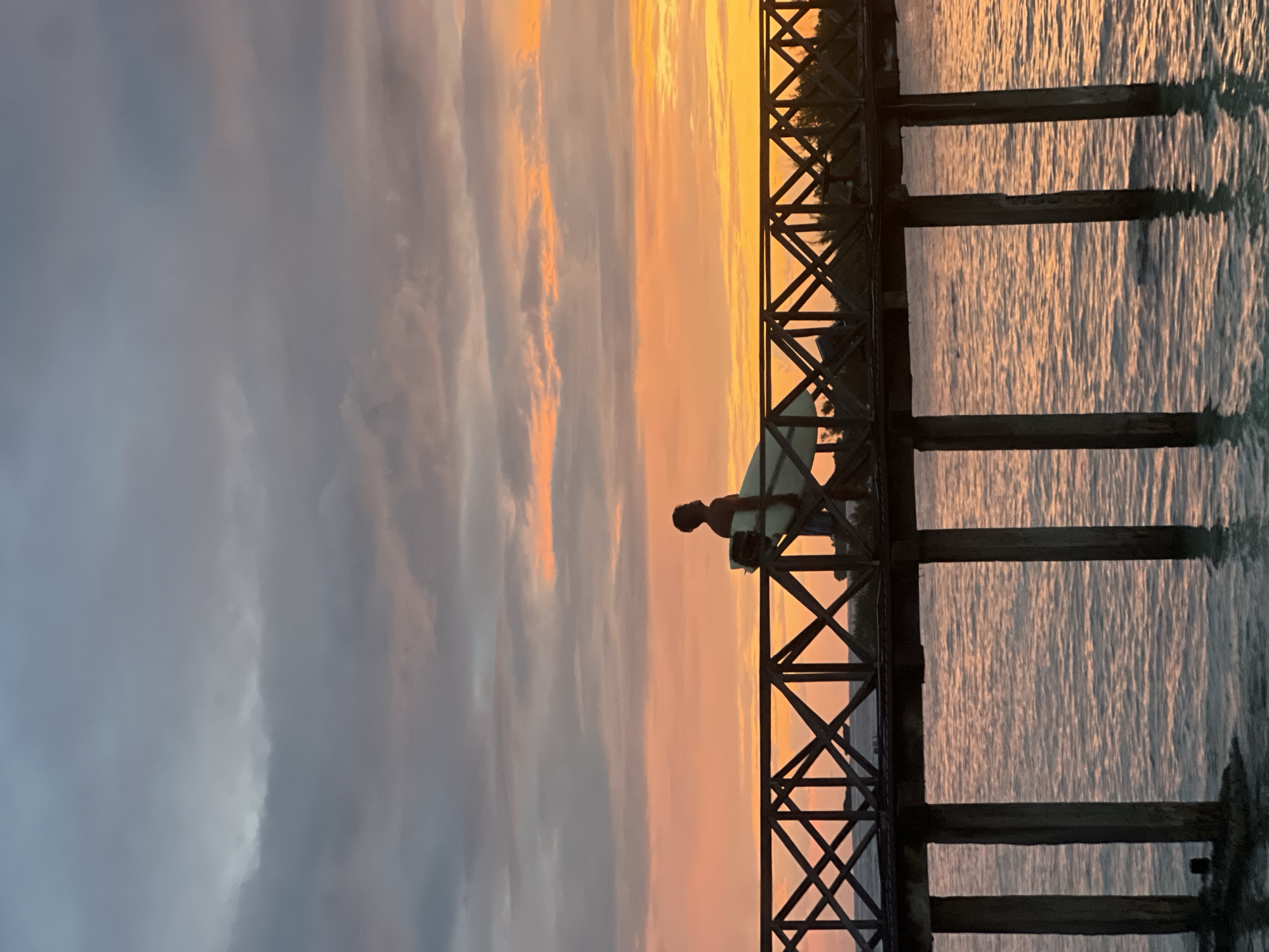 A lone surfer, silhouetted against a vibrant sunset, walks across a wooden pier in General Luna, Philippines. The sky is a canvas of warm oranges and pinks, with streaks of gray clouds overhead. The pier, constructed with dark wooden planks and cross-braced railings, extends over calm, reflective water that mirrors the sunset's colors. The surfer carries a white surfboard, its fins visible, under their arm. The piers' wooden stilts are submerged in the water, showing signs of wear. The overall scene evokes a sense of peaceful solitude at the end of the day.