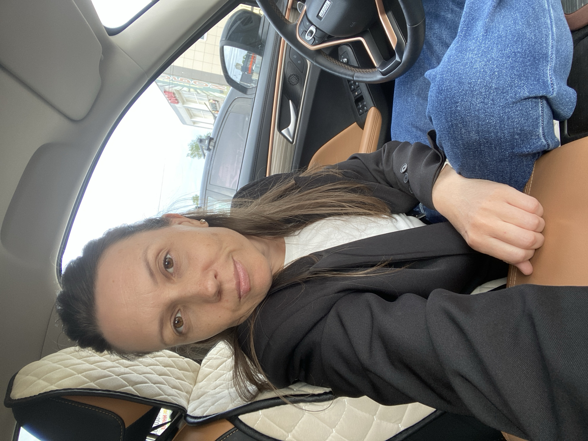 A woman is sitting in the driver's seat of a car. She is wearing a black blazer over a white shirt and blue jeans. The car has a brown interior and a steering wheel with the brand name "Haval." The car seat has a white, quilted seat cover. Outside the car window, buildings are visible, and the signage suggests the location is Chaikovsky, Russia. The woman is facing the camera with a slight smile.