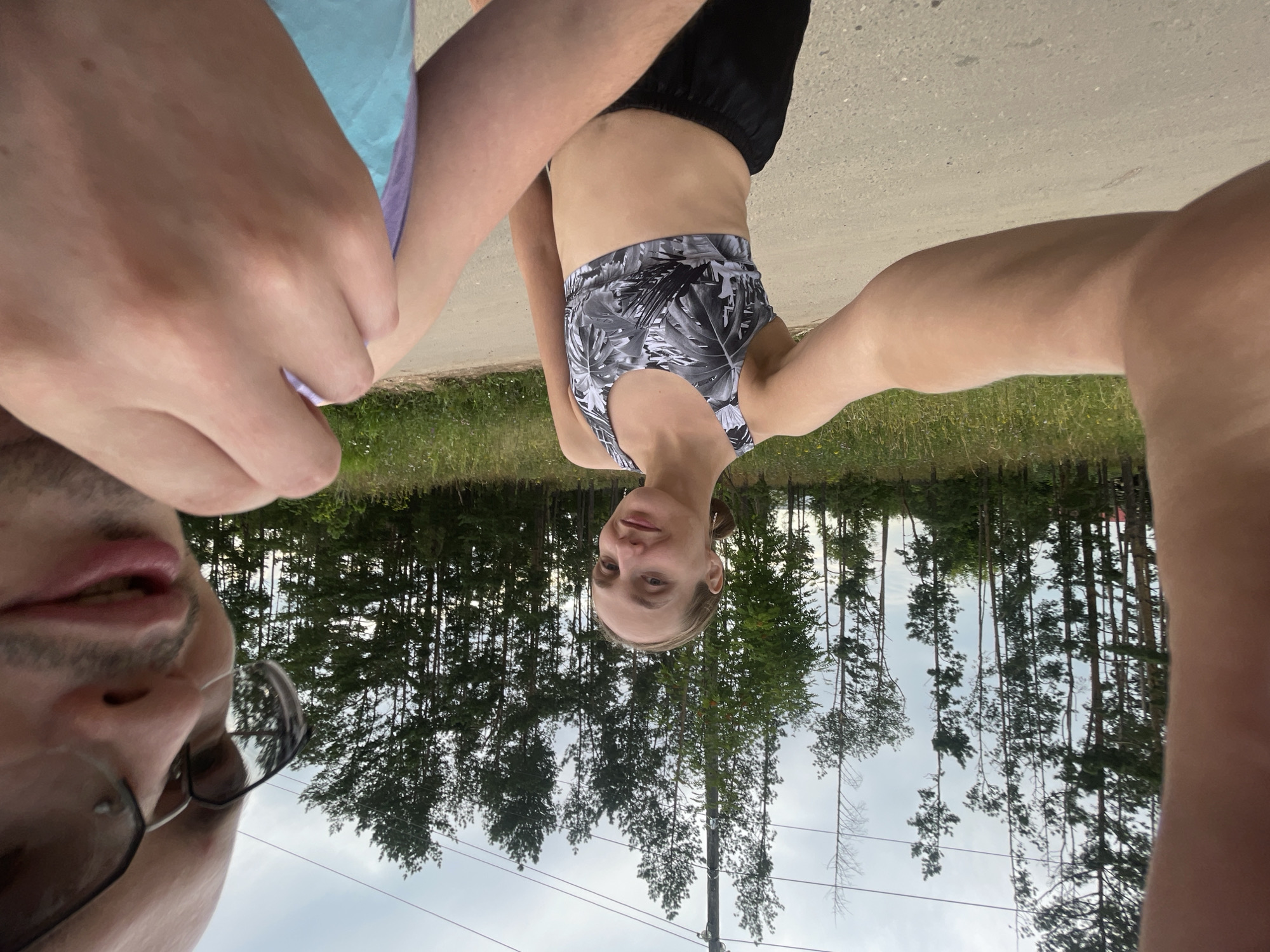 This is a low-angle, upside-down selfie taken outdoors. Two people are visible. On the left is a close-up of a man's face, wearing glasses and a t-shirt. His mouth is slightly open. Next to him, on the right, is a woman wearing a patterned sports bra and black shorts. Her face is visible, and she appears to be smiling slightly. Her arms are extended, and she is holding the camera. They are standing on a dirt path, with a grassy area and a dense forest of pine trees in the background. The sky is overcast. The scene suggests a casual outdoor activity, possibly a walk or hike. The image is oriented upside down, which is a notable detail. There is no visible text in the image. The location context is Podshivalovo, Russia.