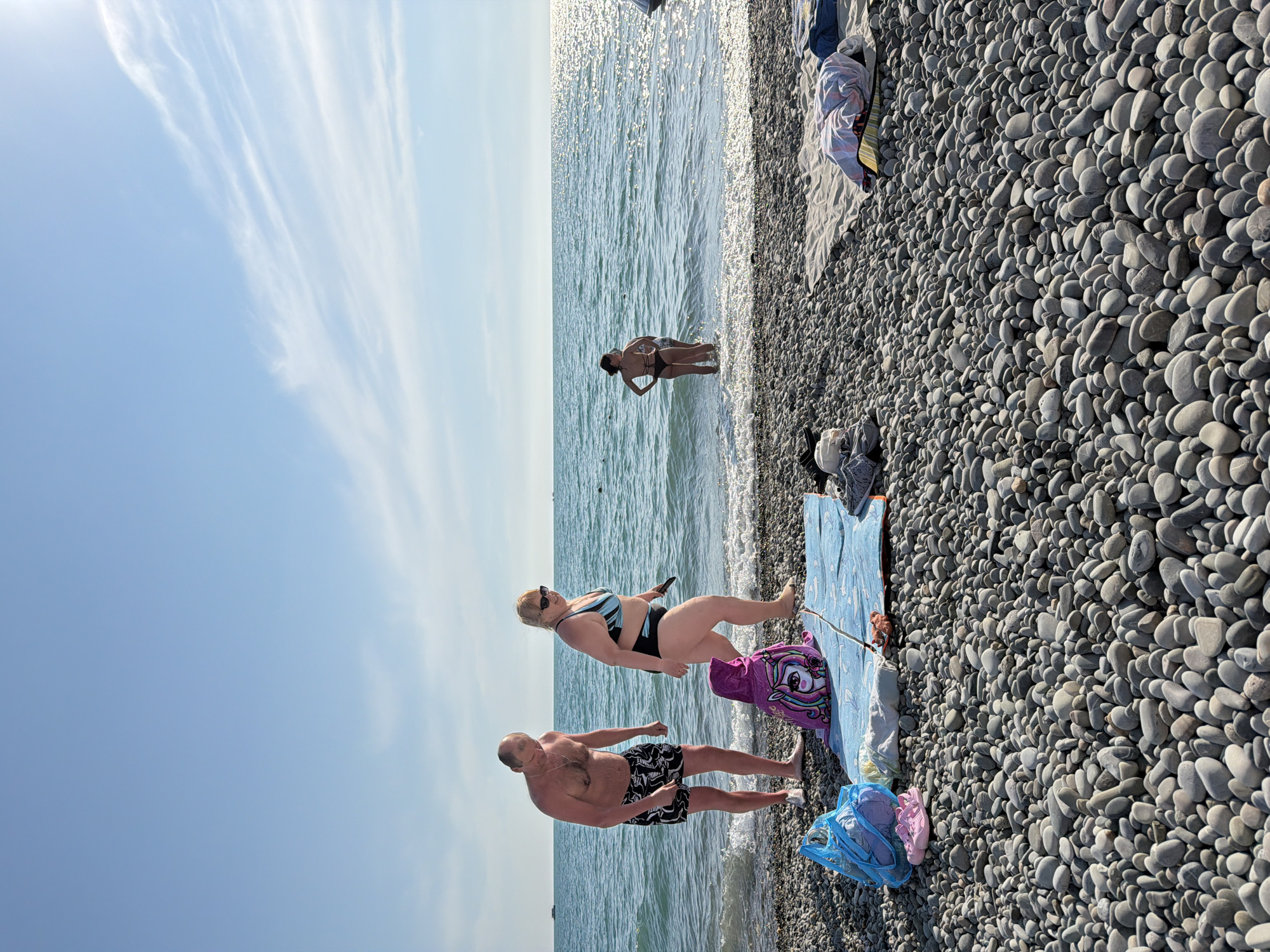 A clear day at Vishnevka beach in Russia features a group of people enjoying the water and shoreline. In the foreground, a man and a woman are standing on the pebble beach, with the man wearing patterned swim trunks and the woman in a blue and black bikini. A child's poncho with a unicorn design is laid out on a blue beach towel between them. Behind them, two women are wading in the shallow, sparkling blue water. The sea stretches to the horizon under a bright blue sky with wispy white clouds. Beach gear, including a blue mesh bag with pink sandals and other items, are visible on the pebbles.