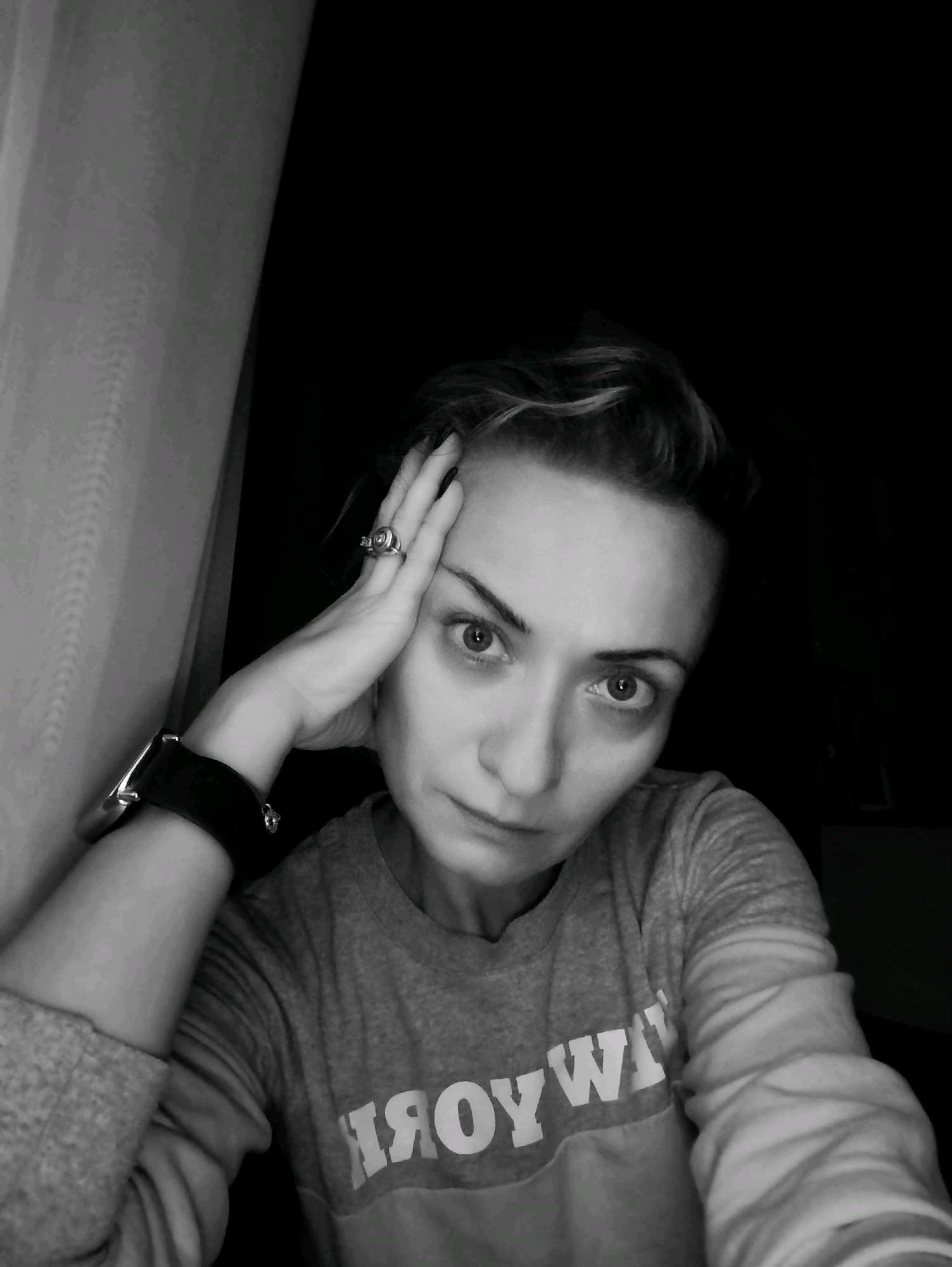 A black and white, medium close-up shot shows a woman with short, styled hair, resting her right hand on her forehead. She is looking directly at the camera with a serious expression. She is wearing a grey long-sleeved shirt with white lettering across the chest that appears to read "YOYWI". On her left wrist, she wears a wide, black bracelet, possibly a smartwatch. On her right hand, she wears a ring with a circular design. The background is dark, with a textured curtain visible to the left of the woman. The image appears to be taken indoors. There is no visible information to indicate the location in Moscow, Russia.