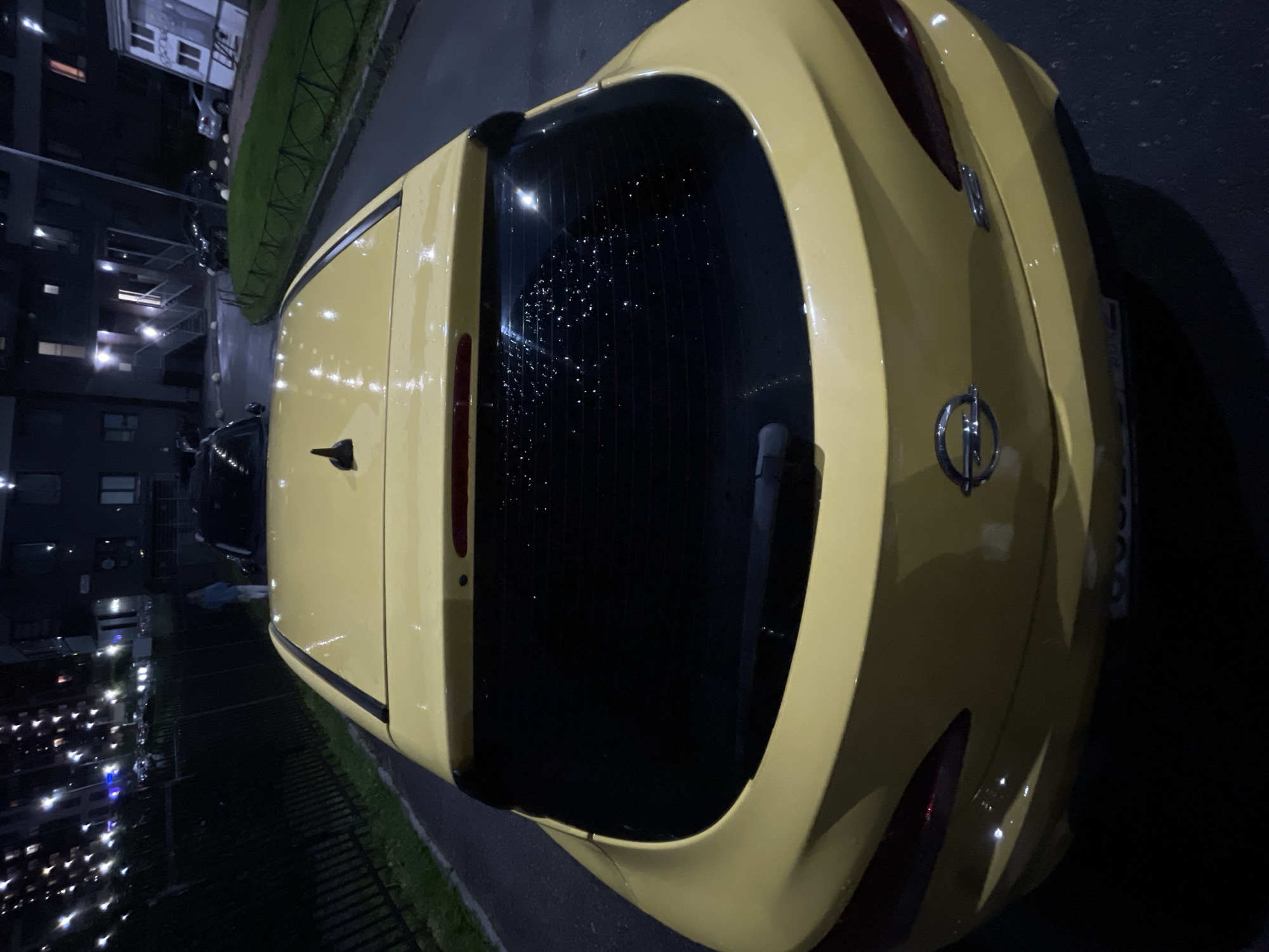 An overhead, close-up, medium shot captures the rear of a bright yellow hatchback automobile parked on a wet asphalt surface at night. The car's rear window is visibly speckled with water droplets, suggesting recent rain. A black spoiler extends from the top of the rear hatch. The car's rear bumper features the Opel logo. To the left of the car, a black metal fence runs along the edge of a grassy area. Beyond the fence, a person in a light blue hooded jacket and white pants is walking away from the camera. In the background, several multi-story apartment buildings are illuminated by numerous bright lights, indicating an urban or suburban residential area. The scene is located in Sinyavino, Russia, as indicated by the environment and typical architecture.