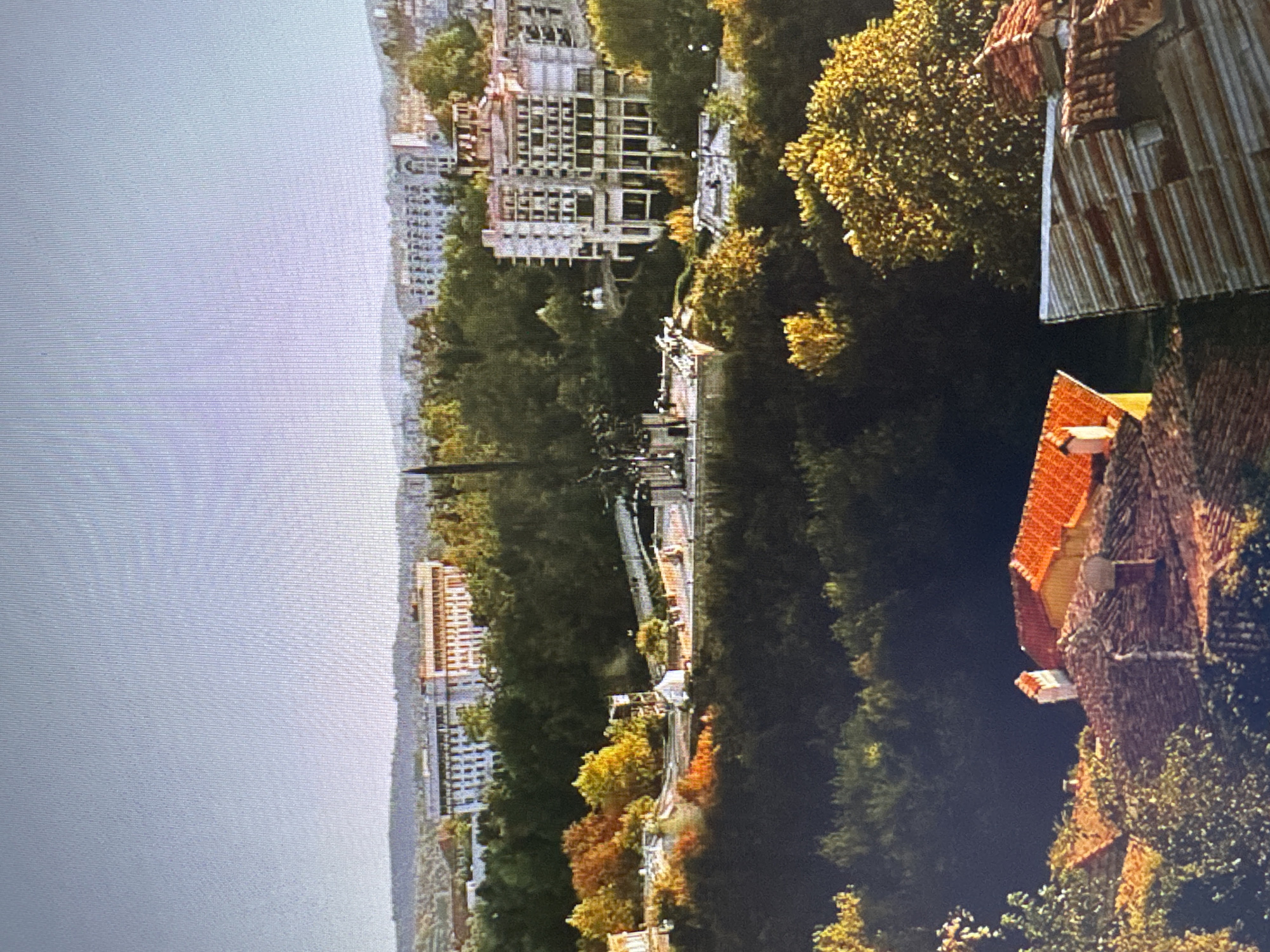 The image depicts a panoramic view of a city, likely Varna, Bulgaria, with a prominent monument and several buildings nestled amongst lush greenery. In the foreground, traditional terracotta-tiled roofs of houses are visible, suggesting an older part of the city. Further into the mid-ground, a large, dark green forested area dominates, interspersed with trees displaying autumnal colors of yellow and orange.
A notable feature in the mid-ground is a monument, possibly the Monument of the Bulgarian-Soviet Friendship or a similar historical landmark, characterized by a tall, dark spire and what appears to be a statue or sculptural elements at its base. The monument stands on a raised platform with paved areas and walkways. Several buildings are scattered throughout the scene. One large, multi-story building with a light-colored facade and numerous windows is situated behind the monument, surrounded by trees. To the right of this, another large building, appearing to be under construction or renovation, with a complex architectural design featuring multiple levels and exposed structural elements, is visible. In the background, more cityscapes with larger, modern buildings can be seen, extending towards a hazy horizon. The sky above is clear and light, suggesting daytime. There are no discernible people or animals in the image. The overall impression is one of a city blending historical architecture, natural landscapes, and modern development.