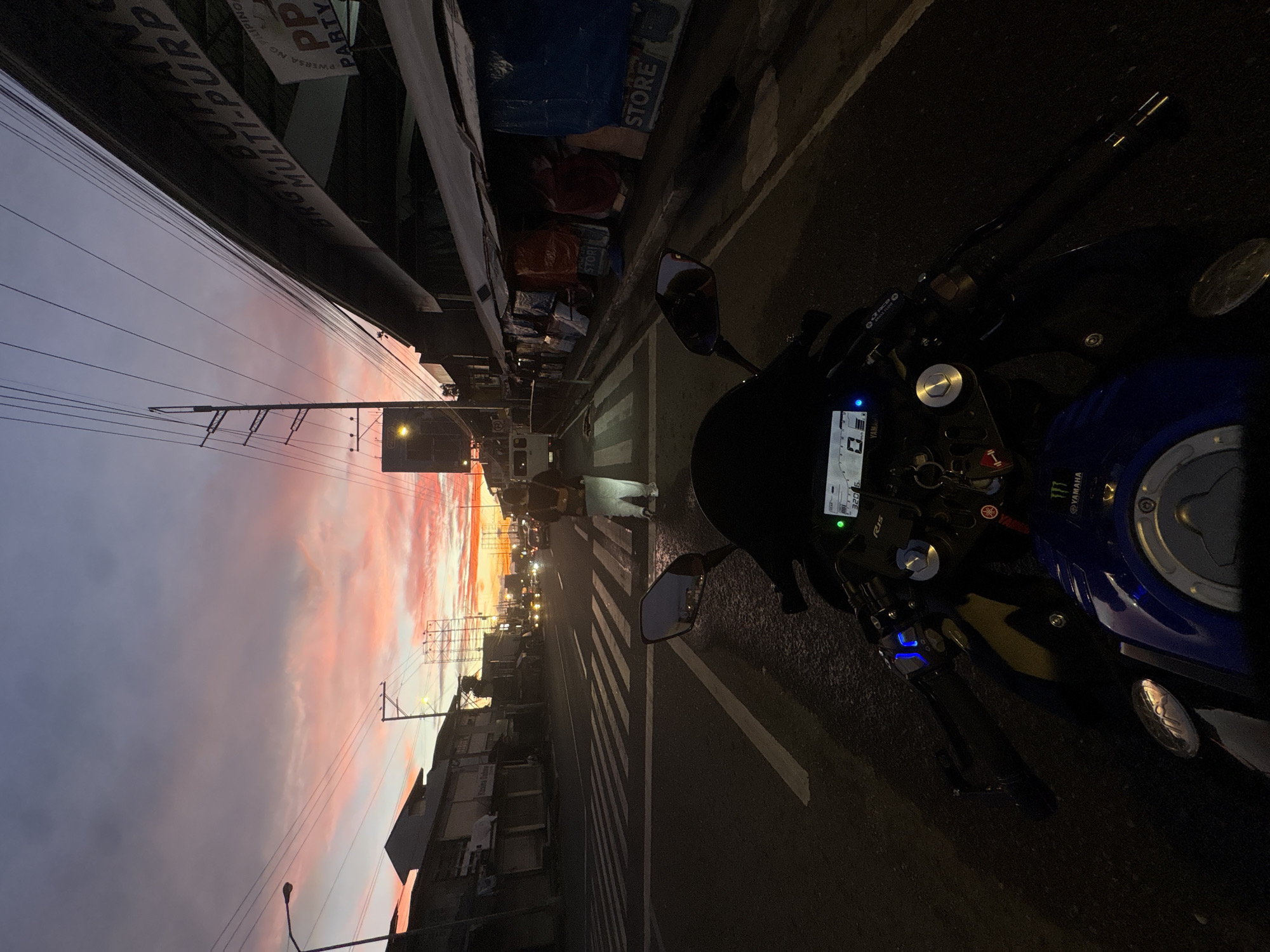 This image depicts a street scene at dusk in Davao City, Philippines, viewed from the perspective of a motorcycle rider. The sky displays a dramatic sunset with hues of pink, orange, and purple, contrasting with the darkening blue. In the foreground, the handlebars and instrument panel of a blue and black motorcycle are visible. The motorcycle has a digital display showing "320".
The scene is a moderately busy street with traffic lights and pedestrian crossing visible. On the left side of the street, there are several buildings with signage that appears to be in Filipino, suggesting a local commercial area. On the right side, a row of stalls or small shops are covered with tarpaulins and displays indicating "STORE". A person wearing a backpack and light-colored pants is standing on the pedestrian crossing in the mid-ground. Various vehicles, including cars and a van, are present on the road, with their headlights illuminated. Overhead, numerous power lines crisscross the sky, a common feature in Philippine urban landscapes. The overall impression is that of a typical evening in a bustling urban environment in the Philippines during the golden hour.