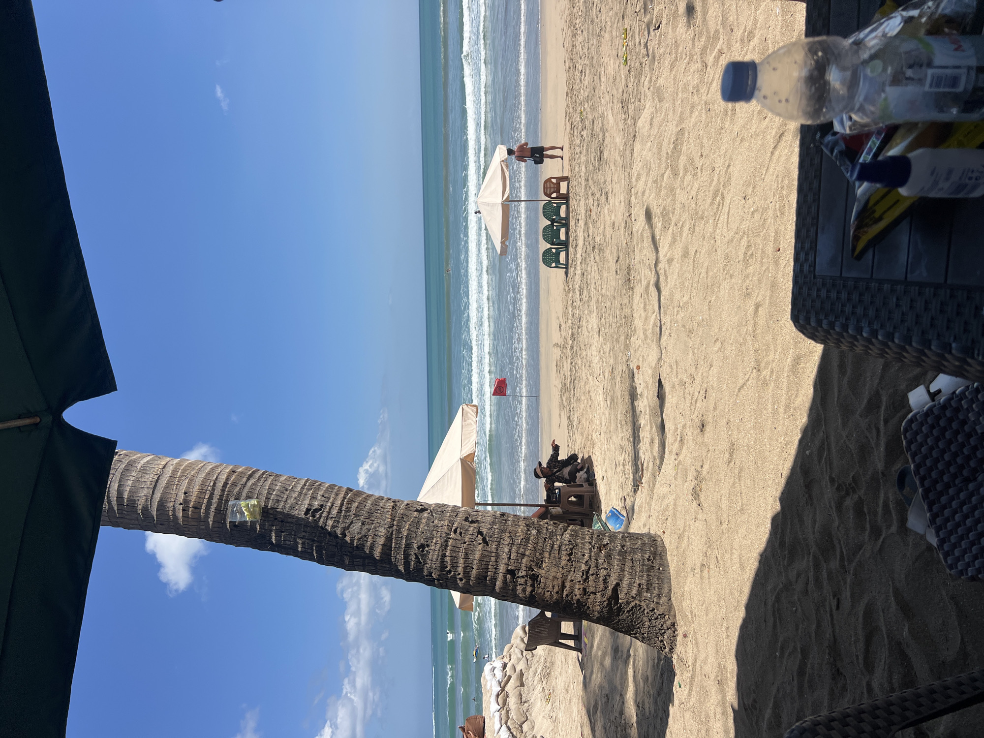 The image depicts a sunny day on Kuta Beach, Indonesia, characterized by a clear blue sky and gentle waves rolling onto a sandy shore. In the foreground, a large palm tree trunk dominates the left side of the frame, partially obscuring the view of the beach and ocean. Underneath a dark umbrella in the foreground, a wicker table holds a plastic water bottle and some other items, suggesting a relaxed setting.
The middle ground of the scene reveals a few people enjoying the beach. Under a light-colored umbrella, two individuals are seated on chairs, with one person appearing to be interacting with the other. A red flag is planted in the sand nearby. Further down the beach, another umbrella is set up with several chairs arranged around it. A man stands near this umbrella, facing the ocean, perhaps a lifeguard or beach attendant. In the background, several surfers can be seen in the water, indicating typical beach activities. Sandbags are stacked to the left, possibly for coastal protection. The overall environment is a typical beach scene in Kuta, with elements of relaxation, leisure, and aquatic activities.