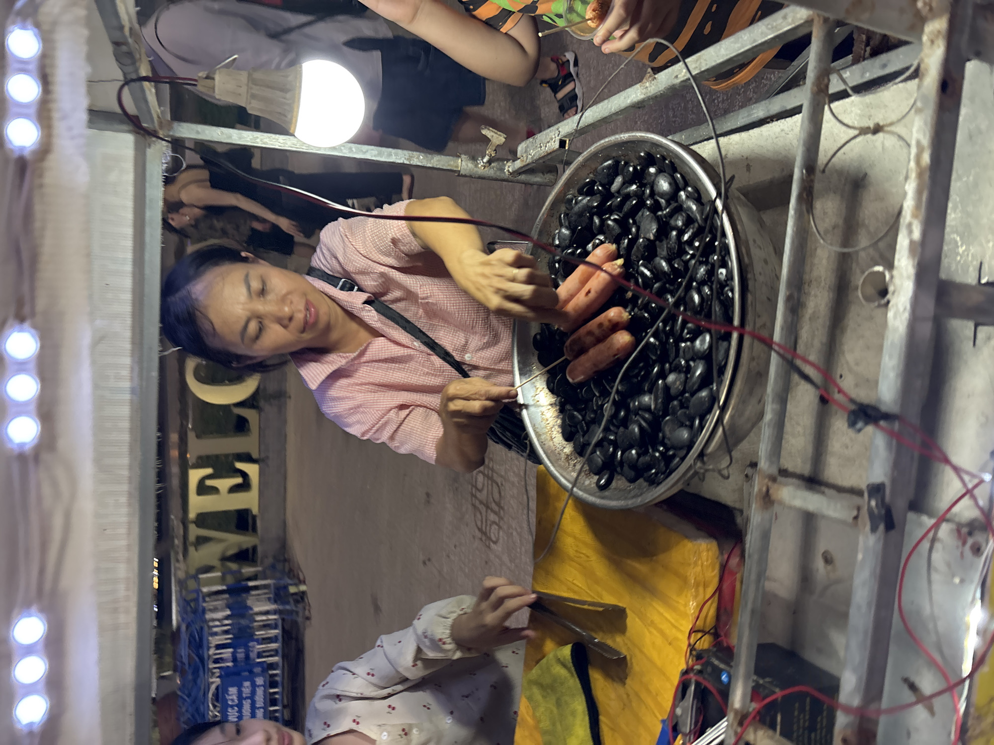 A street vendor is grilling sausages over black rocks at a night market in Nha Trang Ward, Vietnam. The vendor, a woman with short black hair, wears a pink and white checkered shirt and is using tongs to turn the sausages in a large metal bowl filled with black rocks. A younger girl in a patterned shirt is visible to the left, and another person in an orange and black striped shirt is to the right, holding a small cup. Behind them, a large "WELCOME" sign with illuminated letters is visible. The scene appears to be outdoors, possibly on a sidewalk or street, with a metal cart and wires connected to a light bulb overhead.