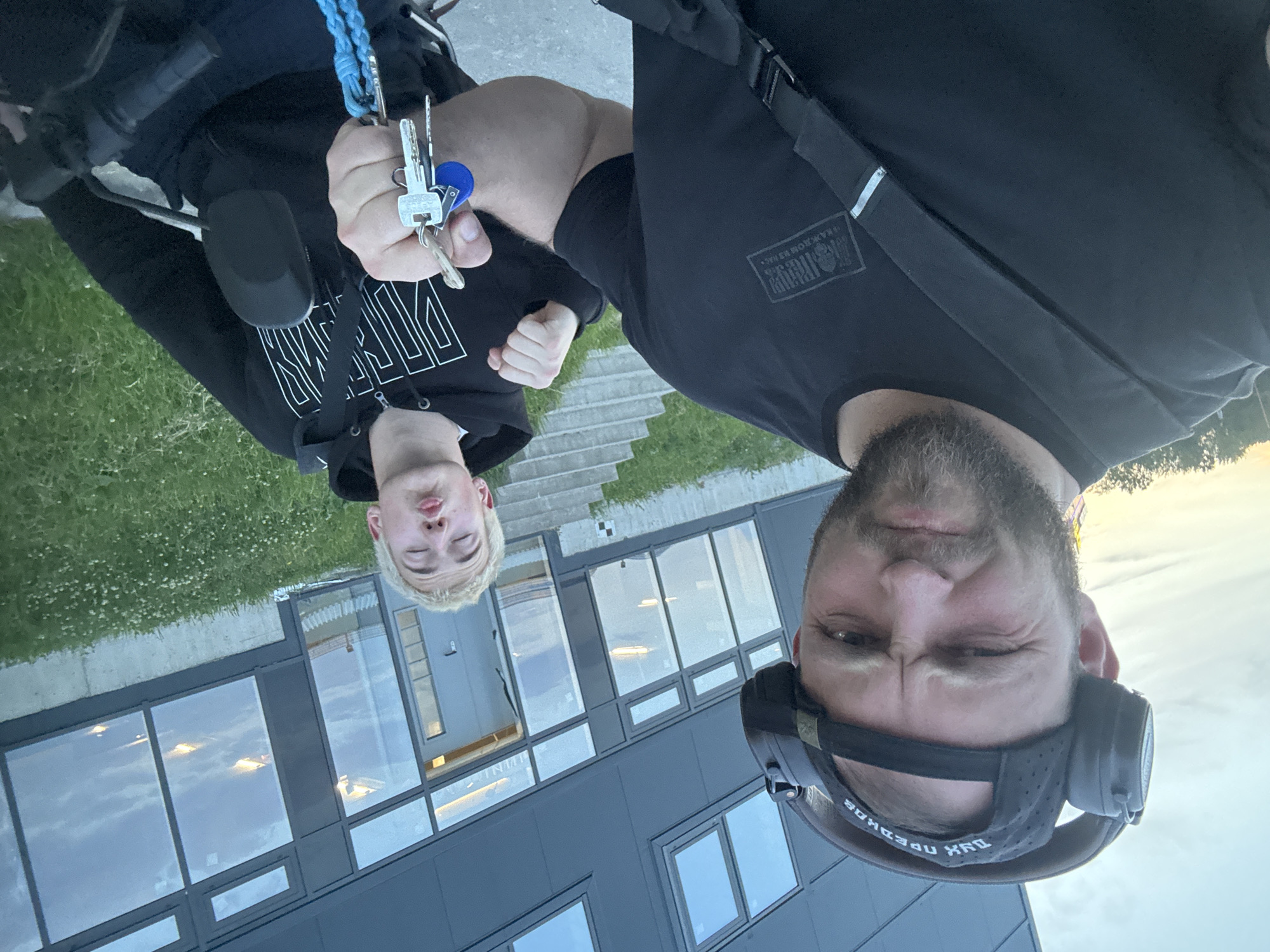 Two men are in the foreground of the image, posing for a selfie. The man on the left is wearing headphones and a black baseball cap with text on it, and a black t-shirt. He is looking directly at the camera with a scrunched-up expression. The man on the right is wearing a black hoodie and is puckering his lips towards the camera. He is holding a set of keys in his right hand. They appear to be on a paved area in front of a modern building with large windows. The building has grey siding and a concrete foundation. There are stairs leading up to an entrance on the right side of the building. Green grass and scattered white flowers are visible in the background. The sky is overcast with hints of sunset.