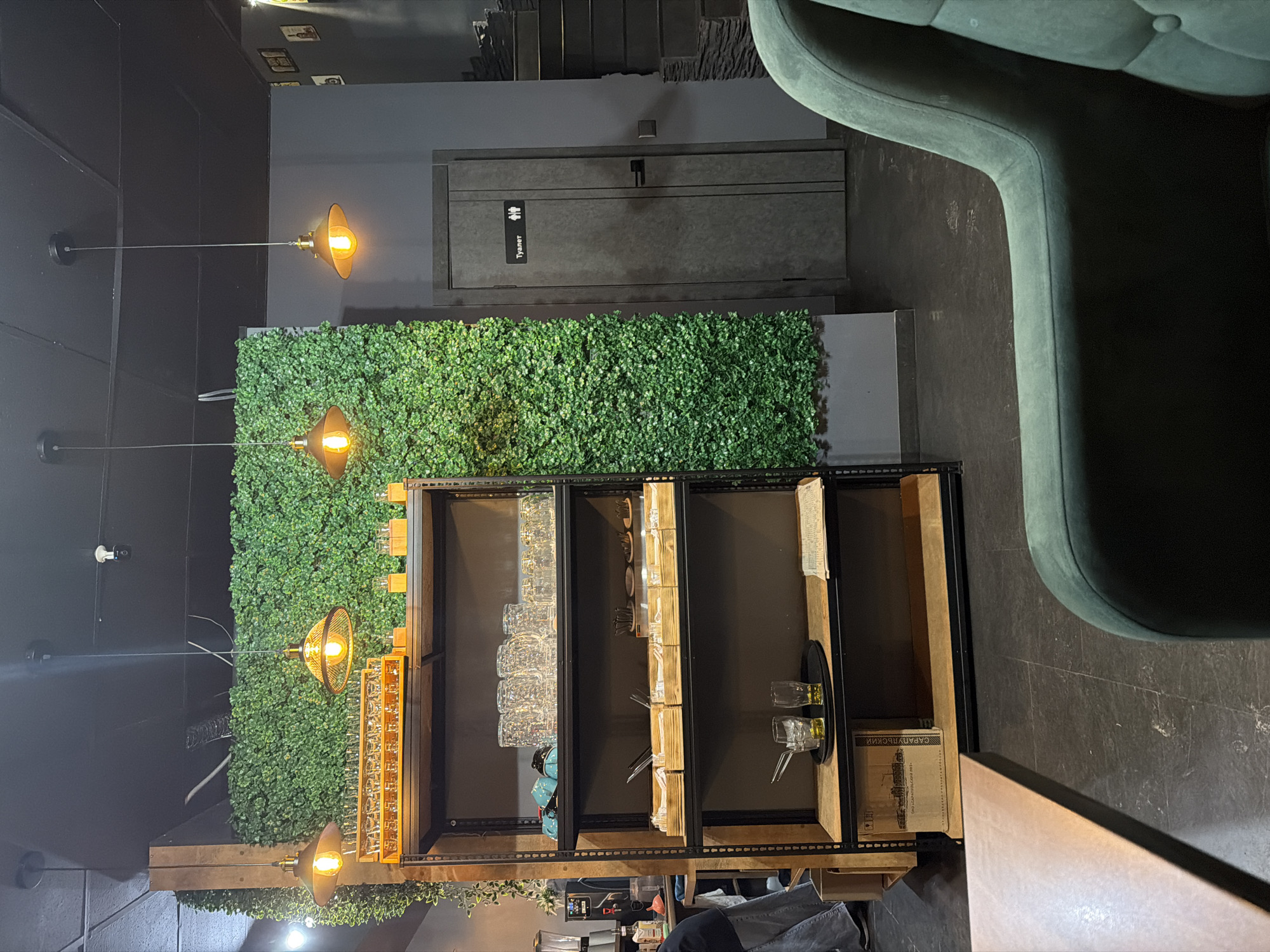 The image depicts an interior space, possibly a restaurant or bar, with a focus on the wall decor and furnishings. A shelf unit, stocked with glassware and tableware, stands in front of a wall covered in artificial greenery. Above, industrial-style pendant lights illuminate the scene. Adjacent to the greenery is a dark gray wall with a door labeled "туалет" indicating restrooms in Russian. A section of a green upholstered chair is visible on the right side of the frame. A portion of a table is visible in the lower right of the scene, indicating a seating arrangement. A partial view of a person near some supplies is in the bottom center of the scene. Given the "туалет" sign and the presence of the supplies, the location is likely in Sarapul, Russia.