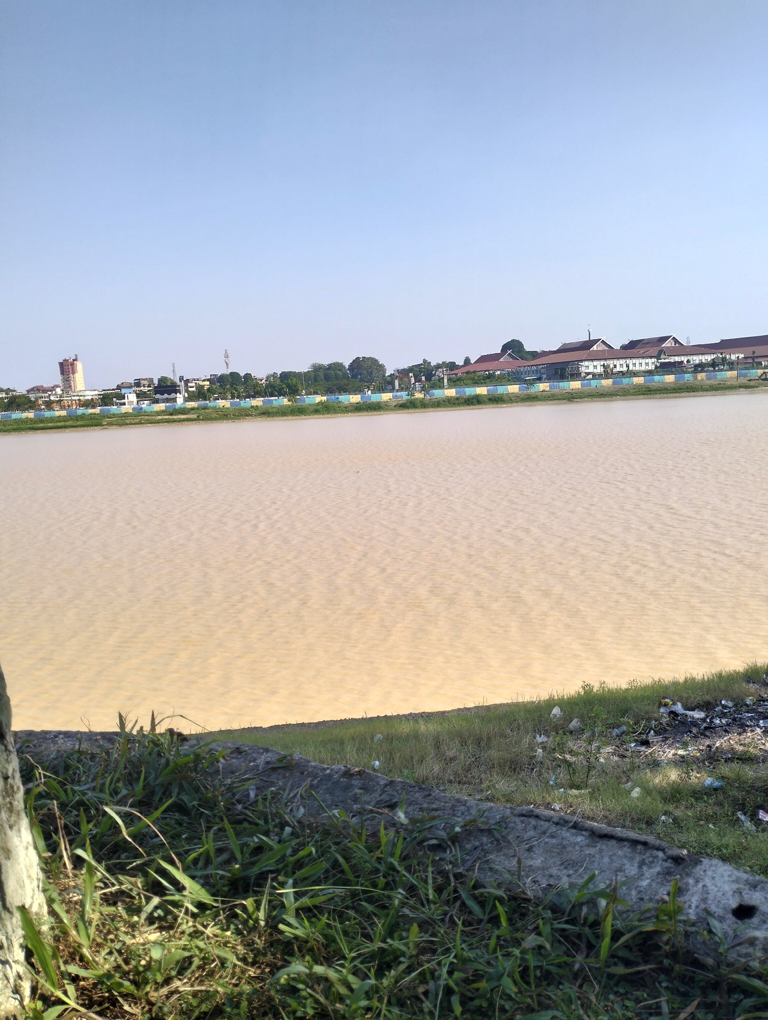 The image depicts a wide, muddy body of water with visible ripples on its surface, suggesting it is a lake or a slow-moving river. The water's color is a light brown or tan, indicative of sediment. Along the far bank of the water, a line of buildings and trees is visible. The buildings appear to be a mix of residential and possibly commercial structures, with some having reddish-brown roofs. A tall, slender tower, possibly a water tower or communication tower, stands to the left of the buildings. The sky above is clear and pale blue, indicating a bright day. In the foreground, the edge of the water meets a grassy bank with some concrete structures and scattered debris, including plastic bags and other litter. The vegetation in the foreground is dense, with tall blades of grass and some weeds. The overall scene suggests an outdoor location within a city, likely Jambi, Indonesia, given the architectural styles and the presence of a body of water, which are characteristic of the region.