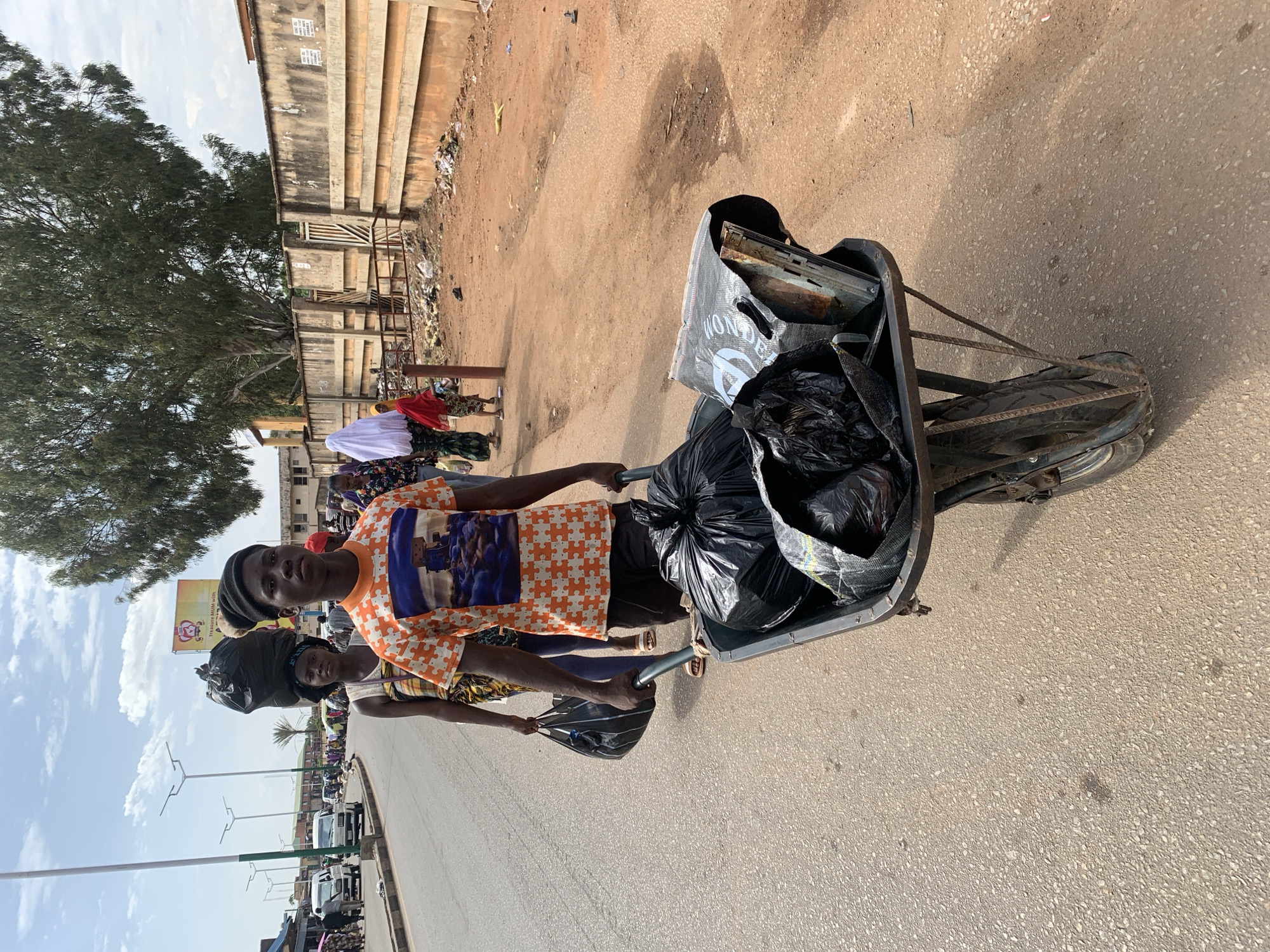 Two individuals are pulling a wheelbarrow filled with garbage along a paved road. The wheelbarrow contains black garbage bags, miscellaneous trash, and a canvas-like bag. The person in the foreground is wearing an orange and white patterned shirt, while the other person is wearing a colorful garment and headwrap. The scene appears to be in Jos, Nigeria, based on visual clues like clothing, terrain, and structures in the background. There are buildings, trees, and parked vehicles visible in the background. Other people are present in the scene. There is visible trash and debris along the side of the road in one area.
