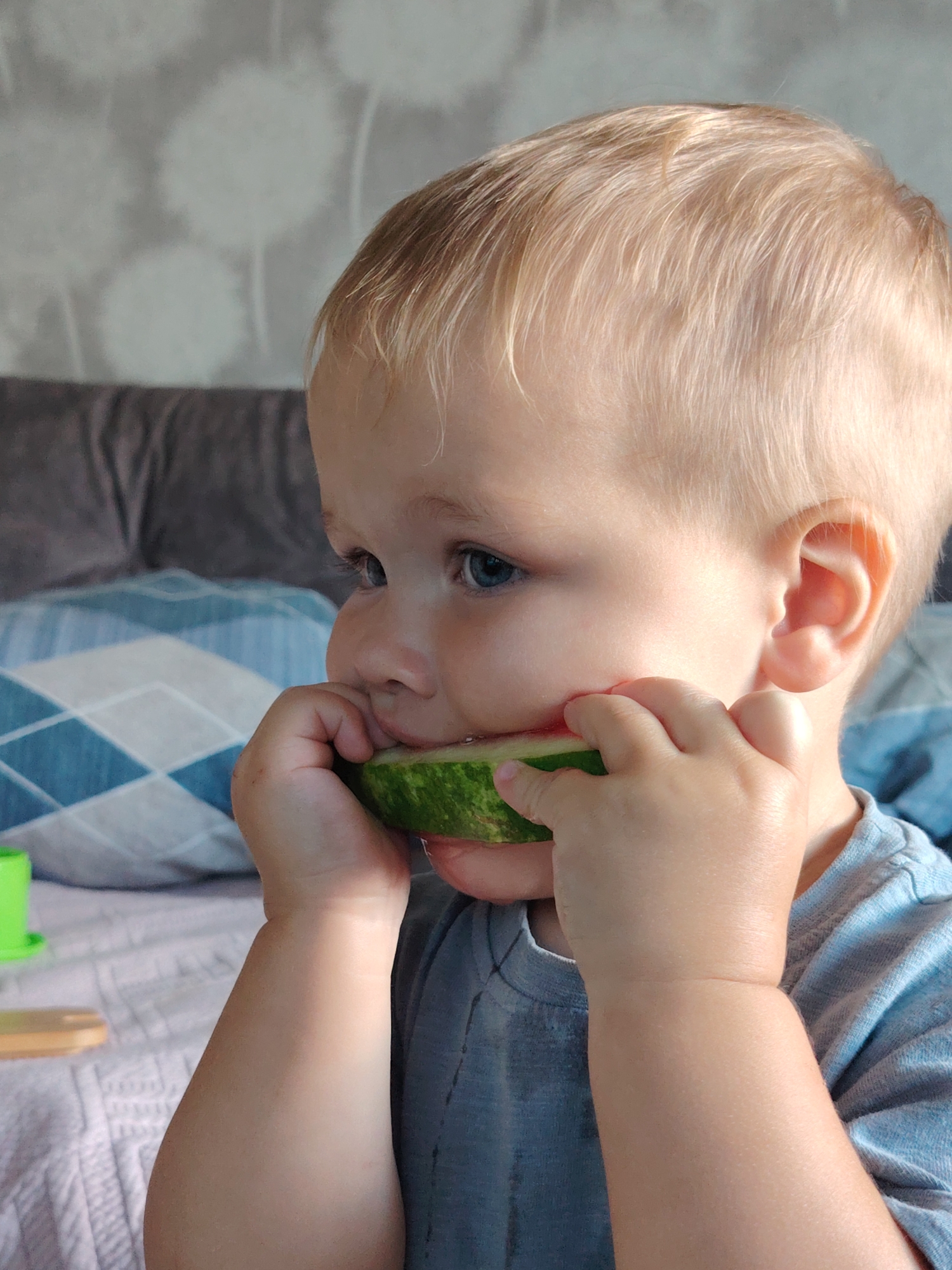 In this close-up image, a young child with fair hair and blue eyes is seen in an indoor setting, likely a bedroom. The child is holding a slice of watermelon to their mouth, with their hands supporting the fruit. The background shows a bed with pillows, one of which has a blue and white diamond pattern. The wall behind the bed has a textured wallpaper with a subtle floral or dandelion-like design. The lighting appears to be natural, possibly from a window. The overall impression is of a candid moment of a child enjoying a snack. The location of Chaikovsky, Russia, is an external detail not visually present in the image itself.