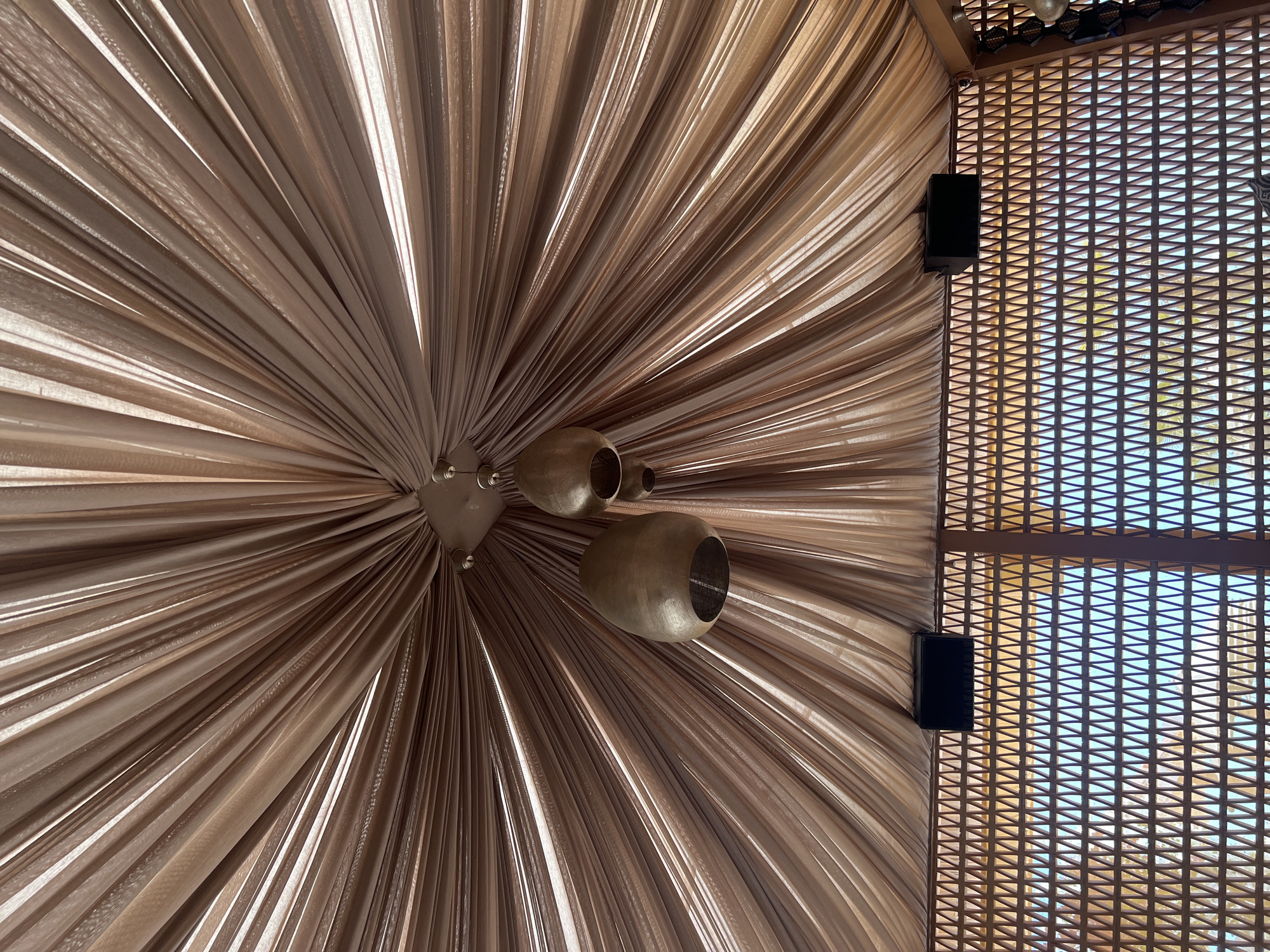 The image depicts the interior of a structure, possibly an outdoor dining area or pavilion, with a draped ceiling and decorative light fixtures. The ceiling is made of a sheer, light brown fabric that is gathered and suspended from a central point, creating a radial pattern. Two bell-shaped pendant lights, with a metallic bronze finish, hang from this central fixture. Below the draped ceiling, there is a decorative wall or screen with a geometric pattern of triangular openings, through which a bright, possibly outdoor, environment is visible. Two black speakers are mounted on the structure above this patterned wall. The scene is static, with no people or discernible actions. The overall impression is one of an aesthetically designed space, likely intended for relaxation or entertainment. The context of Nesebar, Bulgaria, suggests this could be a venue located in a coastal or historical area of the town.