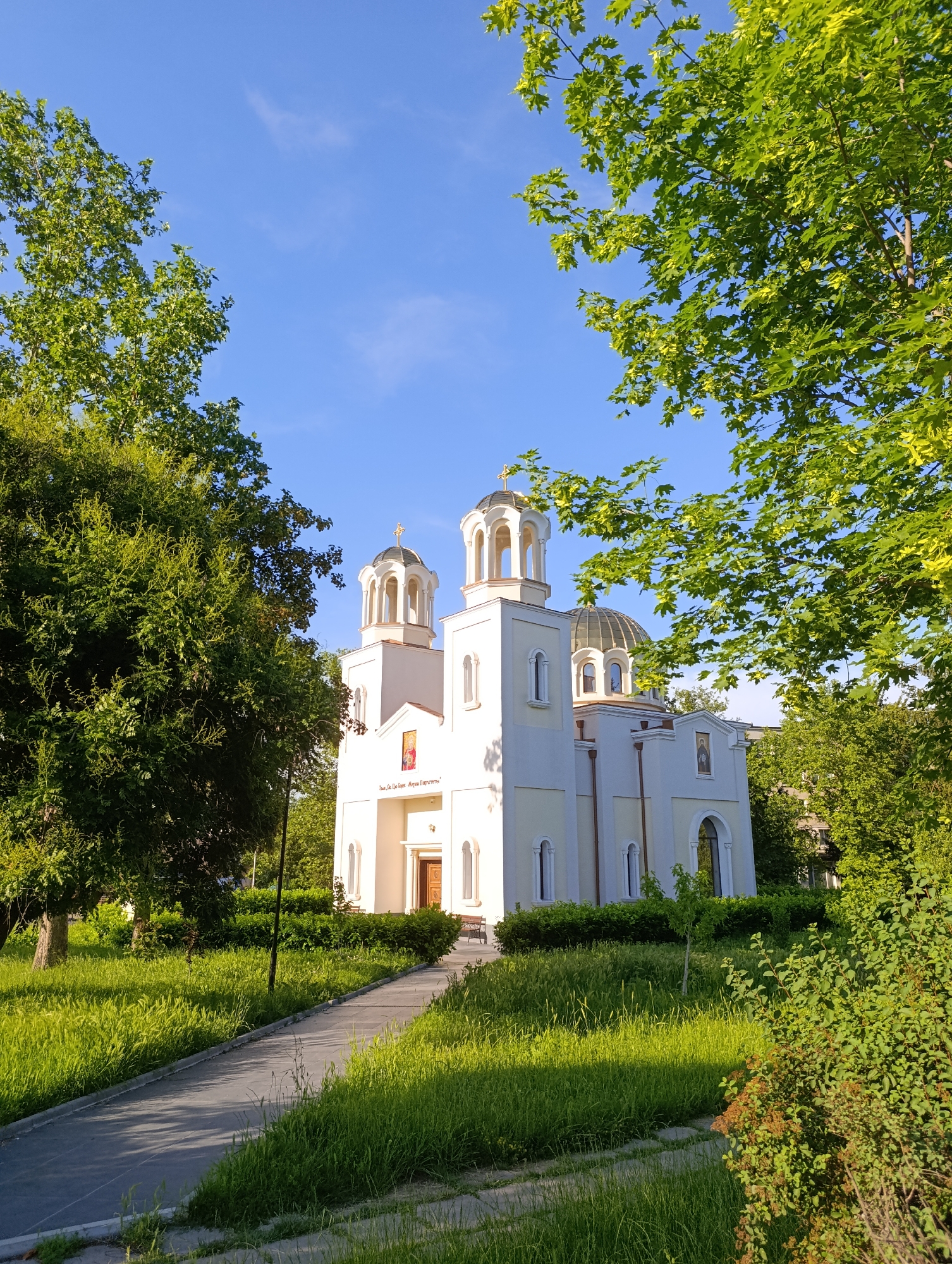 The image shows a white Orthodox church with a golden dome and steeple, surrounded by lush green trees and grass. A paved walkway leads to the entrance of the church. The sky is a clear blue with a few wispy clouds. The scene is bathed in soft sunlight, suggesting it is either early morning or late afternoon. The church appears to be in a well-maintained park or garden. The inscription above the entrance to the church reads "Св. Цар Борис – Михаил". This is the Church of Saint Tsar Boris – Michael in Razgrad, Bulgaria.