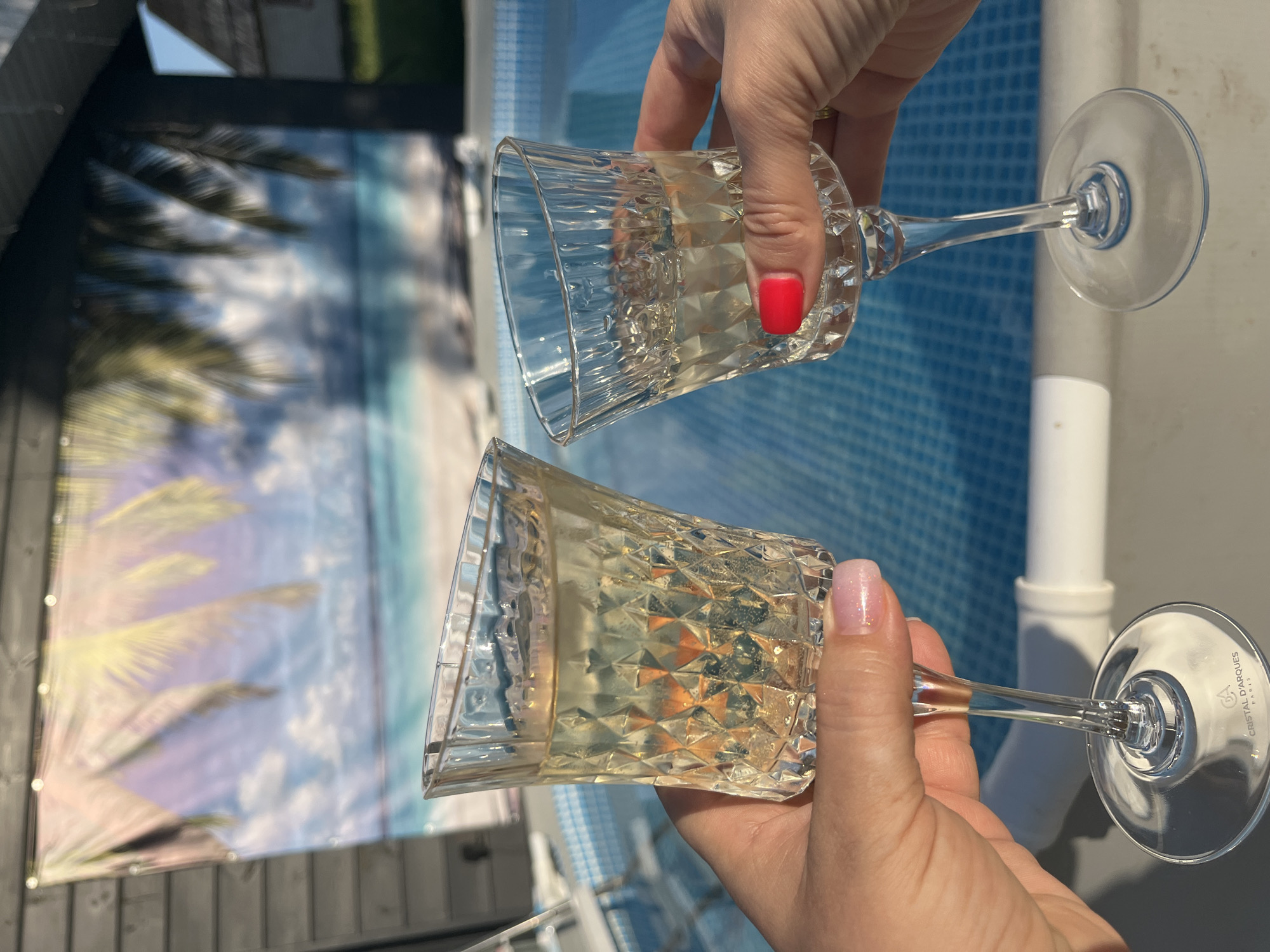 Two hands hold crystal glasses filled with champagne or sparkling wine. The backdrop includes a banner depicting a beach scene with palm trees and the ocean, as well as the edge of a pool. The hands belong to two different people, one with red nail polish and the other with light pink nail polish. The location is inferred to be Новая Чернушка, Russia from the information available in the request. The glasses are branded 'Cristal d'Arques PARIS'. The scene is outdoors.