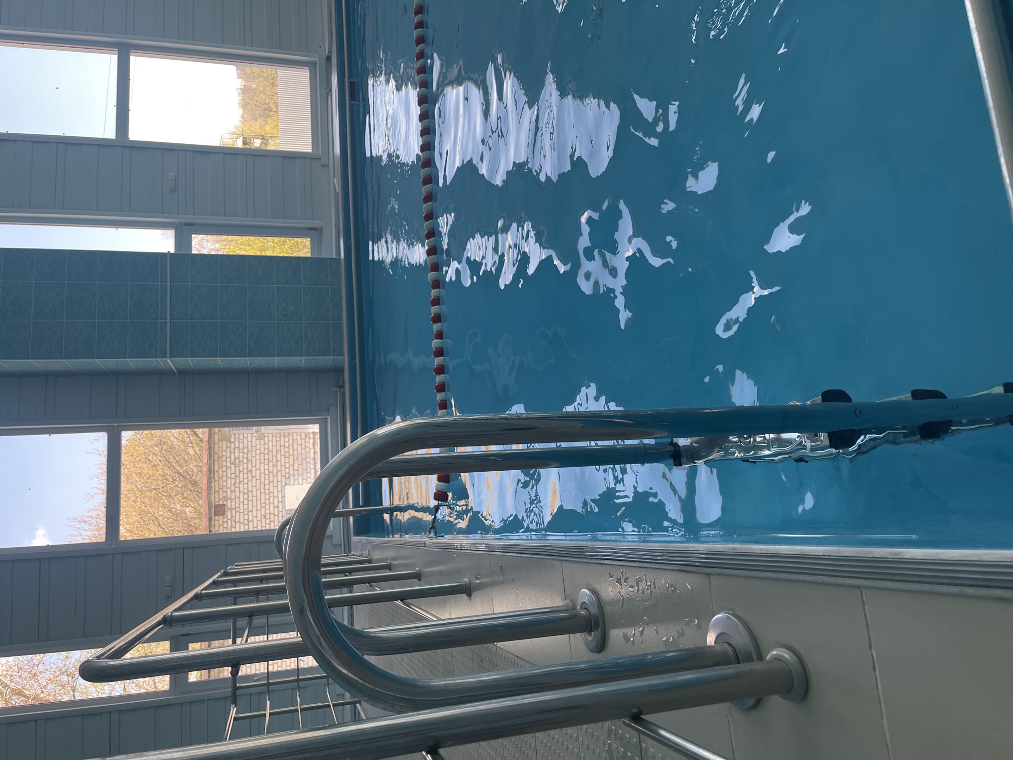 The image depicts the interior of a swimming pool facility in Ulyanovsk, Russia. The view is from the edge of the pool, looking across the water towards the far side. The pool water is a bright blue, with ripples and reflections on its surface. A lane divider marked with red and white floats stretches across the pool. A chrome pool ladder is visible in the foreground. Windows are located above the pool, offering a view of the exterior, including buildings and trees. The walls surrounding the pool are tiled in light blue. The overall environment is clean and well-maintained.