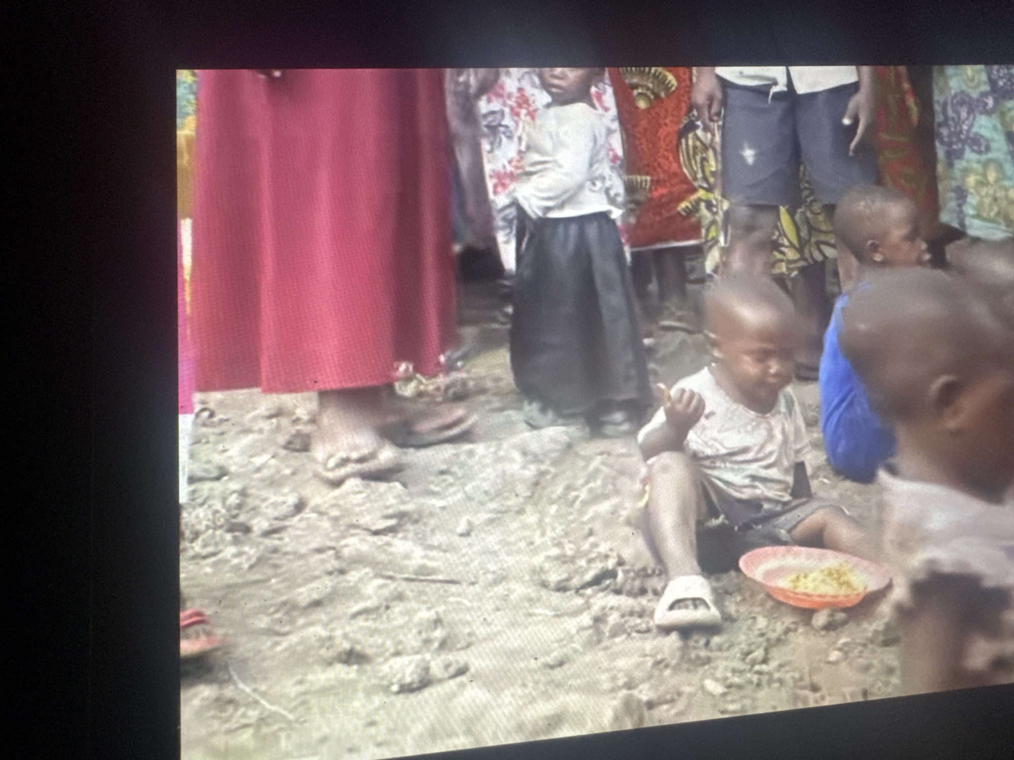 The image depicts a group of people, including children, in what appears to be an outdoor setting. The ground is uneven and appears to be dirt or mud. Some individuals are seated or lying down, while others are standing. There are colorful fabrics visible, suggesting clothing or coverings. One child is holding a shallow bowl containing what looks like food. The overall scene suggests a communal gathering or a moment of rest in a rustic environment. The image is taken from a slightly elevated perspective, looking down onto the scene. There is no clear indication of a specific urban location within Abuja, Nigeria, as the environment appears to be rural or undeveloped.