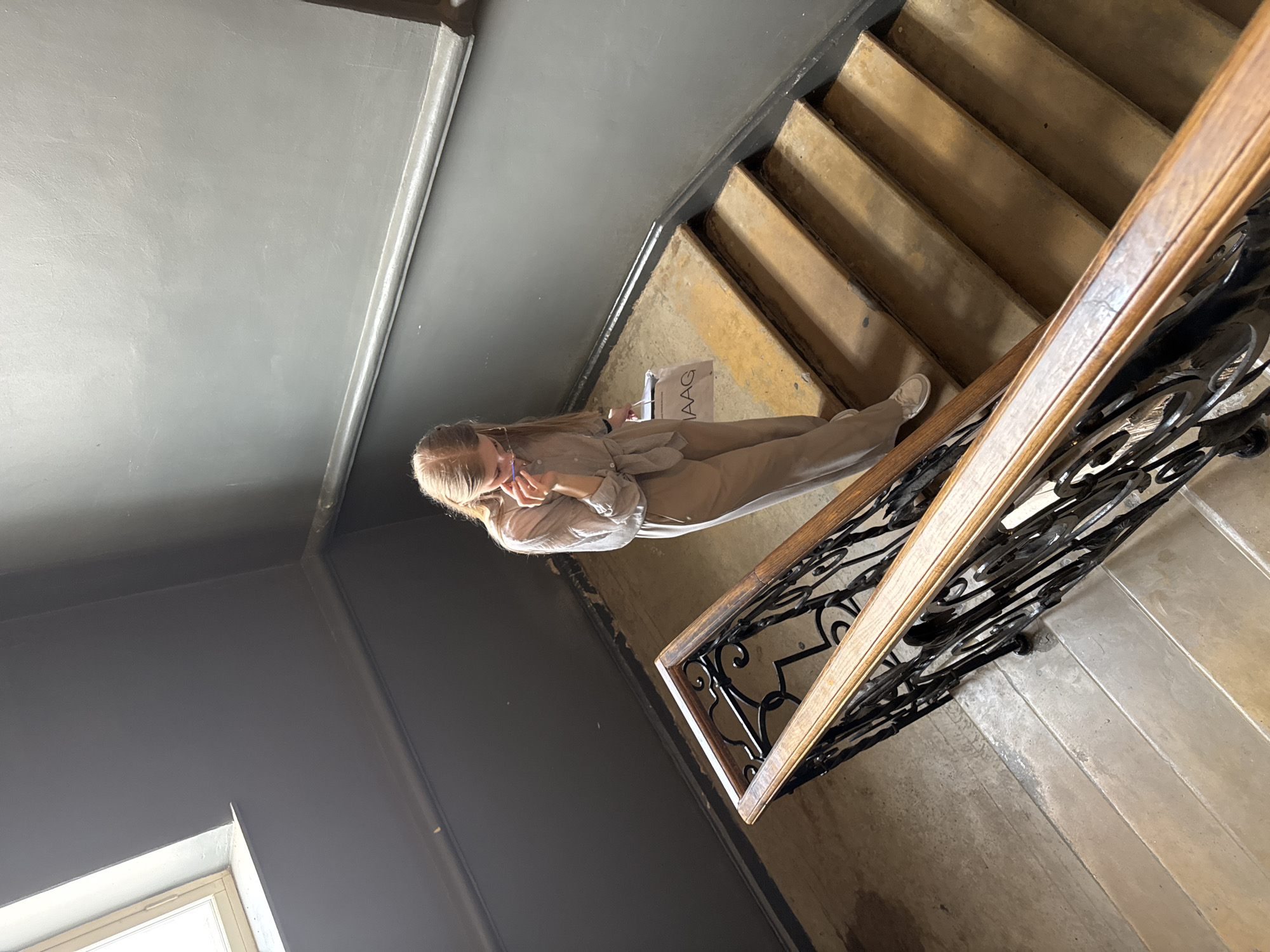 A young woman with blonde hair tied back, wearing a light-colored jumpsuit and white sneakers, walks down a concrete staircase with wooden and wrought-iron railings. She is holding a paper shopping bag with the logo "IAAG" visible. The woman is looking down and touching her face, possibly adjusting glasses. The staircase is located in an interior space, with dark grey walls and a window visible in the upper left corner. The lighting suggests it is daytime. The image does not provide any specific information or visual cues that would allow for identification of the location as being in the city of Perm, Russia.