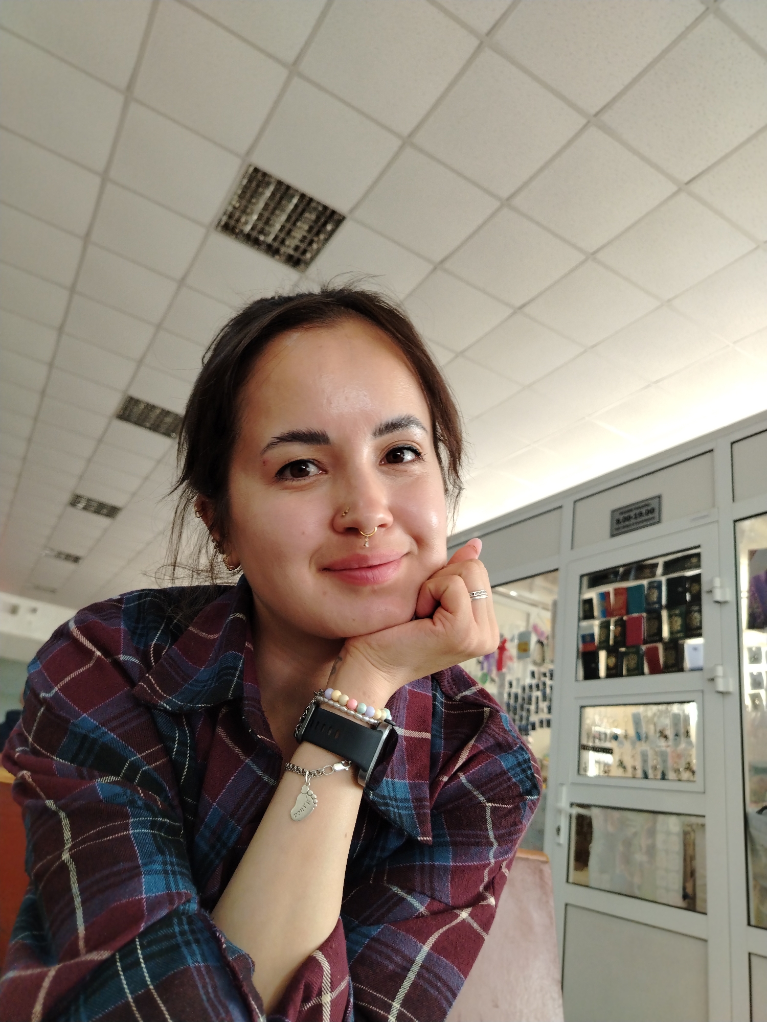 A young woman with medium-length dark hair, a nose piercing, and wearing a burgundy and blue plaid shirt is seated indoors. She is resting her face on her hand, wearing a bracelet with a charm and a smartwatch. The background includes a white tiled ceiling with a recessed light fixture and a white partition with glass panels showcasing various items, possibly for sale. A sign is visible on the partition. It is probable that the location is in the city of Chaikovsky, Russia, but this cannot be confirmed from the image alone.