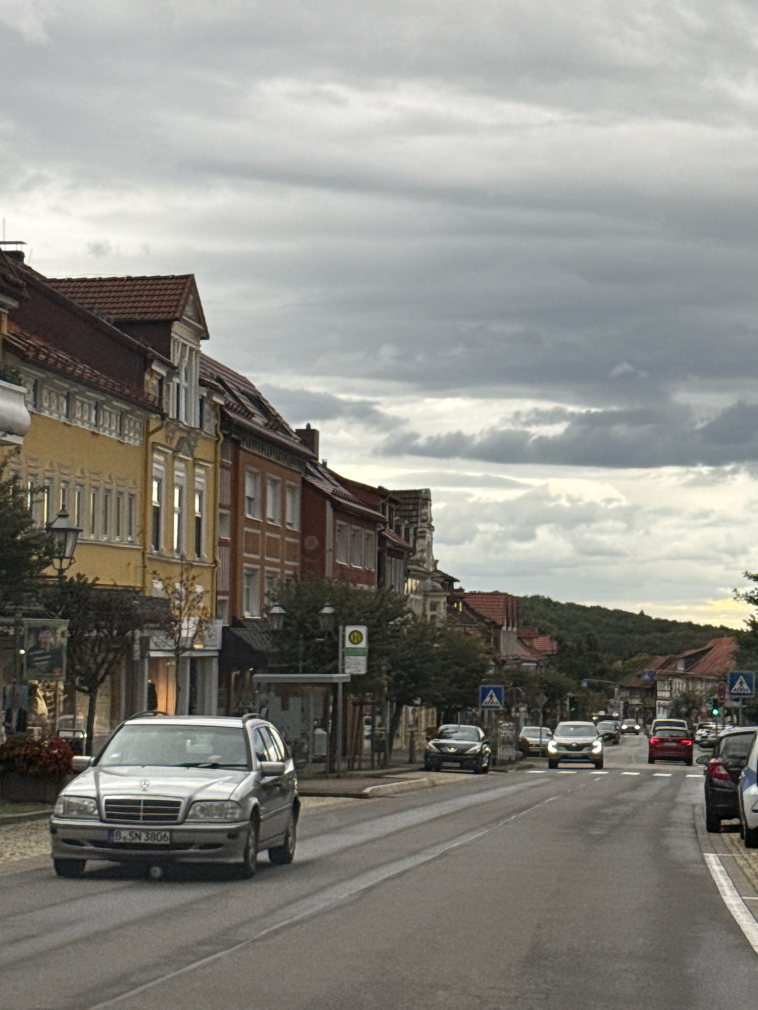The image shows a street scene in Bad Sachsa, Germany. In the foreground, a silver Mercedes-Benz C-Class estate car is driving down the wet asphalt road. The car has a German license plate that reads "B.SN 3806". On the left side of the road, there are several buildings with traditional German architecture, featuring red tiled roofs and colorful facades in yellow and orange. A street lamp with an old-fashioned design is visible in front of these buildings. On the right side of the road, more buildings are present, along with trees and a bus stop with a sign indicating public transport. Further down the street, other cars are seen, including a dark-colored sedan, a red car, and a white car. The sky is overcast with grey clouds. In the background, a densely wooded hill can be seen. The overall atmosphere suggests a somewhat gloomy or autumnal day.