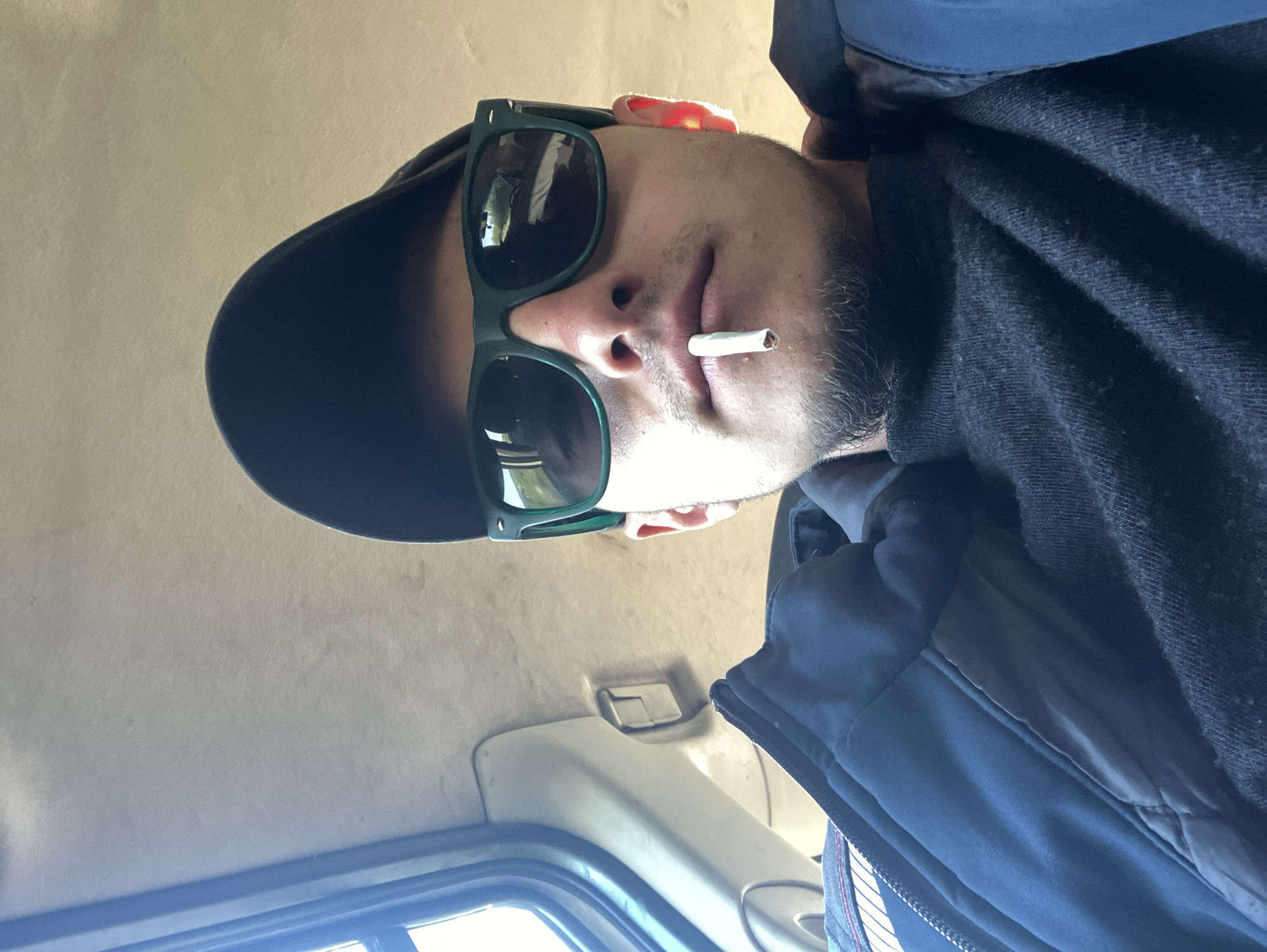 A man is seen from a low angle, wearing a black baseball cap, green sunglasses, and a dark-colored jacket, with a cigarette in his mouth. The setting appears to be the interior of a car, given the overhead interior fixtures and window visible in the background. The image does not provide any information regarding the location in the city of Biaga, Bulgaria.