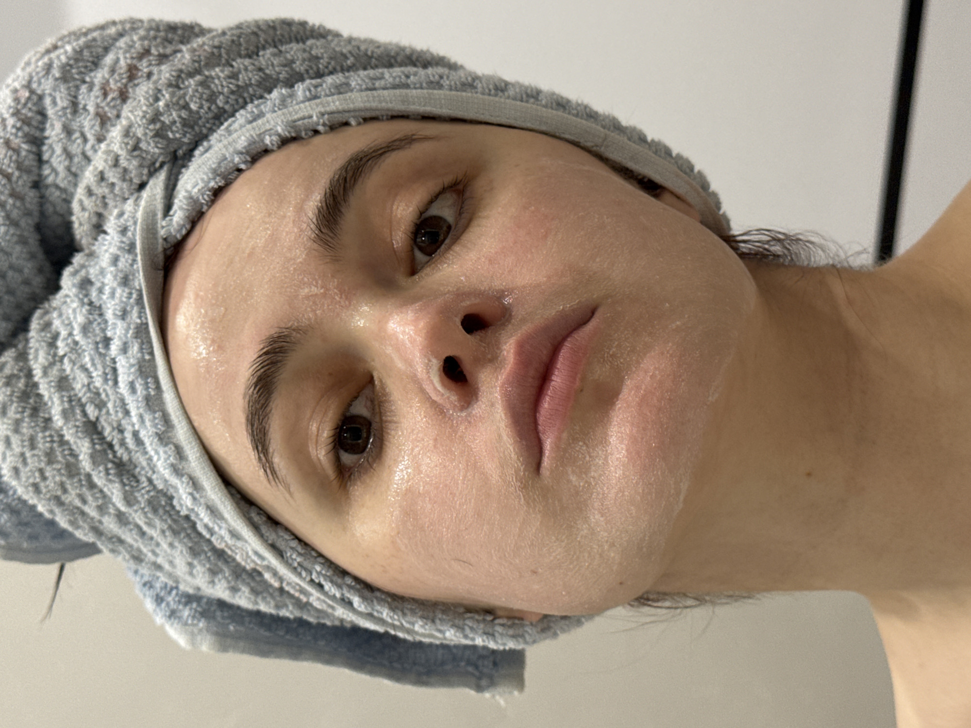 A close-up, high-angle portrait captures an individual's face, tilted, during a skincare routine. The subject, a person with light skin, dark brown eyes, and dark eyebrows, looks directly at the camera with a neutral and focused expression. Their head is wrapped in a light blue or grey terrycloth towel, protecting their hair from the product applied to their face. A white or translucent substance, consistent with a face mask or cleanser, is visibly applied across their facial skin, giving it a somewhat patchy and reflective appearance. The skin underneath the product shows some natural texture. A portion of the person's bare neck or shoulder is visible on the right side of the frame. The background is a plain, light-colored surface, indicating an indoor setting. The lighting is soft and even, illuminating the details of the face and the skincare product. No text is visible.