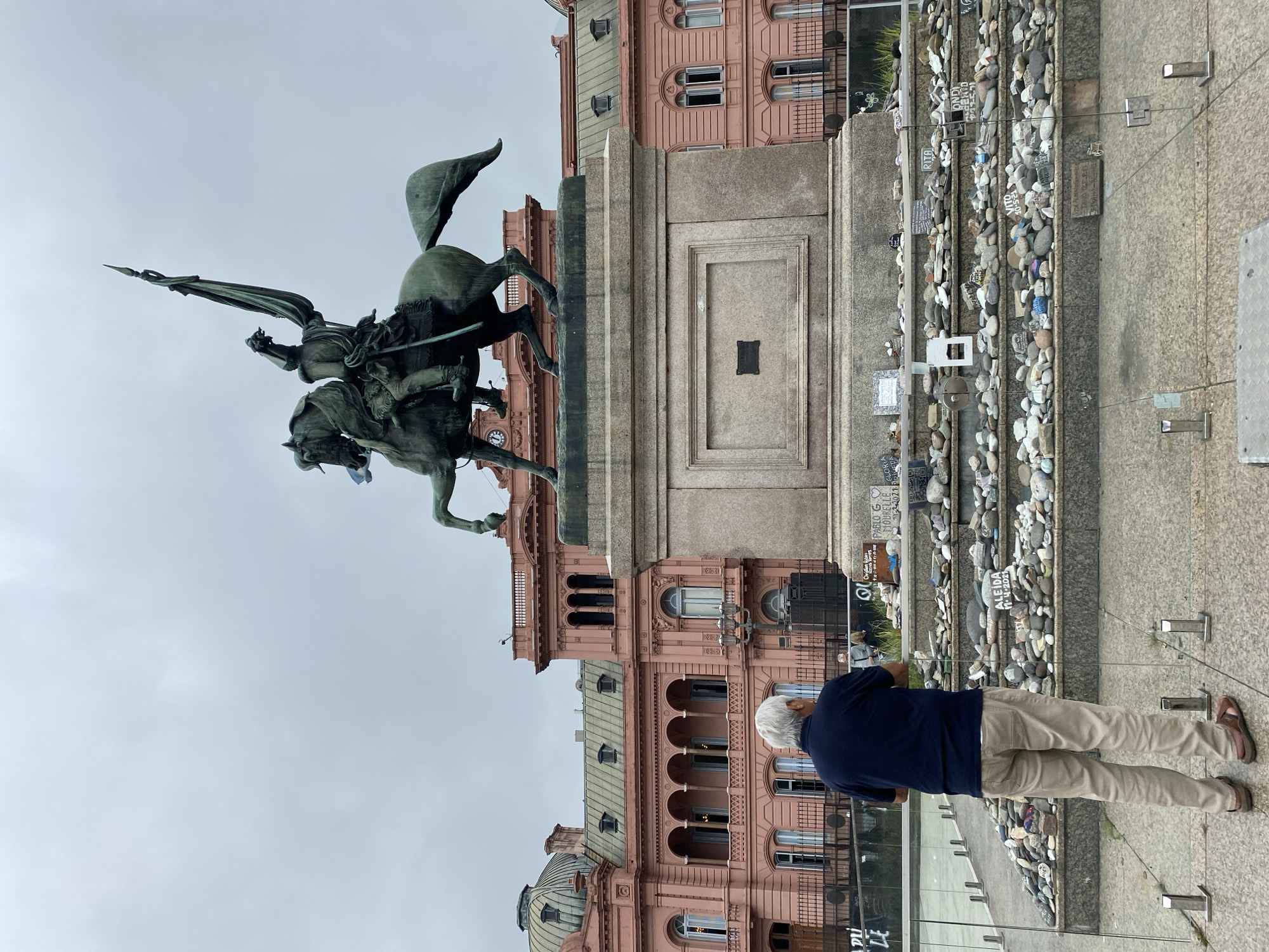 The image captures an overcast day in Buenos Aires, Argentina, featuring an equestrian statue, a historic pink building, and a solemn memorial.
Dominating the center is a large bronze equestrian statue, depicting a male figure in military attire, possibly holding a spear or flag, mounted on a rearing horse. The statue rests on a substantial stone pedestal, which has a rectangular panel with a dark plaque embedded in its center.
Behind the statue stands a grand, ornate pink building, characteristic of the Casa Rosada. It features multiple stories with arched windows, balconies, and decorative elements. A clock face is visible on one of its lower sections. The sky above is uniformly grey, indicating an overcast day.
In the foreground, facing away from the viewer, an older man with white hair is visible. He is wearing a dark blue short-sleeved shirt and light-colored khaki pants, with brown sandals. He stands behind a glass barrier, looking towards the memorial area.
To the right of the statue's base and extending into the foreground is a unique memorial. It consists of multiple tiers or steps covered with numerous small, smooth river stones and plaques. Many of these stones and plaques have handwritten or inscribed names and dates, suggesting a personal tribute site. Visible text on some of these memorial items includes "RITA," "VITO," "ALBIDA 11-4-2022," "GLORIA," and "PABLO G MORBIELLE." This area conveys a sense of remembrance and reflection. The overall scene suggests a significant public square, possibly the Plaza de Mayo, given the prominent pink building.