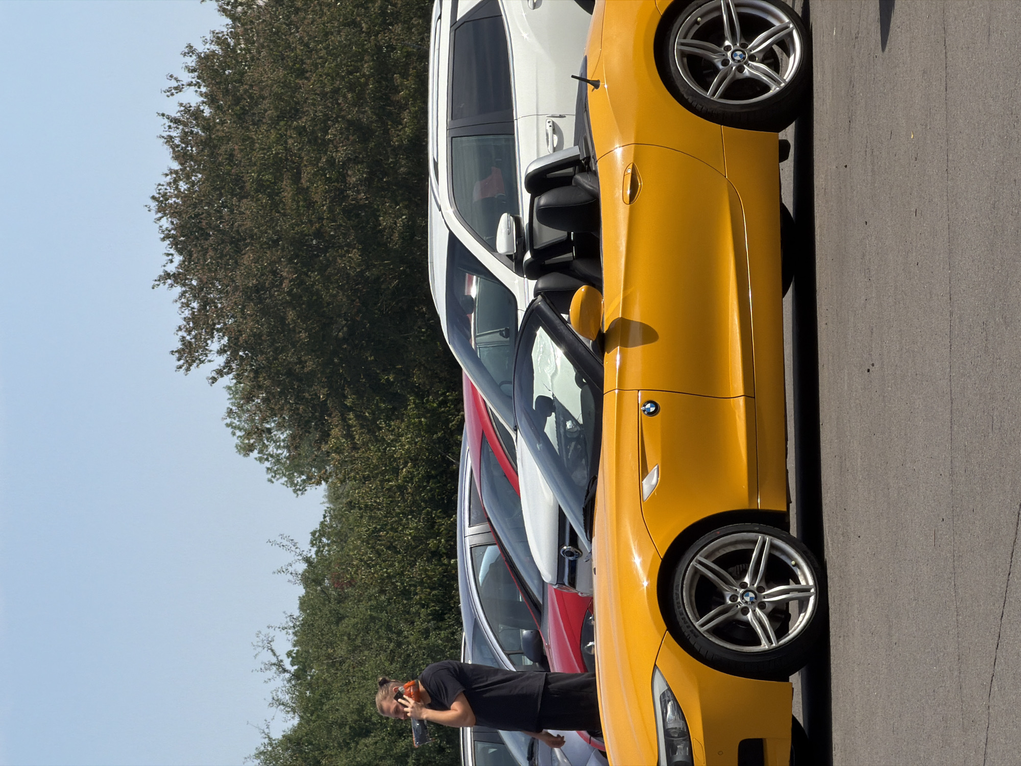 A blonde man with a bun is standing on the left side of the image, wearing a black t-shirt and black shorts, holding a mobile phone to his ear. He is standing next to a line of cars parked in an outdoor lot. In the foreground, a bright yellow convertible BMW Z4 is parked. Behind the BMW, a white SUV and a red car are visible. The background consists of green trees under a clear blue sky. The scene appears to be in Mondercange, Luxembourg, given the context of other images from this location.