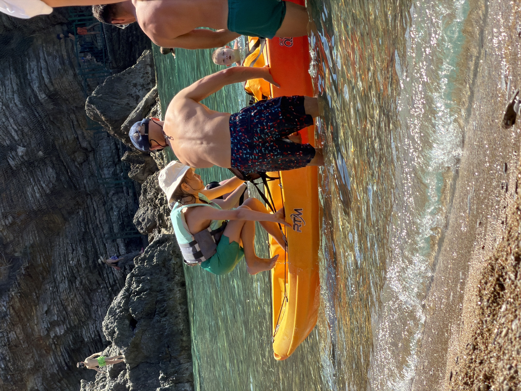 This image depicts a sunny coastal scene near Budva, Montenegro, characterized by large, horizontally stratified grey rock cliffs meeting clear, turquoise water and a narrow, pebbled beach.
In the midground, an orange "RTM Ocean Duo" tandem kayak is positioned in shallow water. A young person, wearing a light green life vest, green swim shorts, and a light-colored hat, is boarding the kayak from the water, with bare feet partially inside. An adult male, viewed from behind, wearing patterned swim shorts and a dark blue baseball cap, stands waist-deep in the water, assisting the boarding process. To his right, another adult male, also viewed from behind and wearing dark green swim shorts, stands in the water. A young child's face and shoulders are visible in the water behind these individuals.
In the background, on the prominent rock formations, a young person in light green swim shorts stands on a rock to the left. To the right, another person in a hat and patterned clothing is walking downwards on the rock face, near a green railing structure. Further back and higher on the cliff, an individual in a dark shirt and red shorts is visible on a path with a green railing system.
The immediate foreground shows shallow, clear water with ripples and small waves breaking on a light brown, coarse-grained sand and pebble beach. The scene is brightly illuminated by direct sunlight, creating highlights on the water's surface.