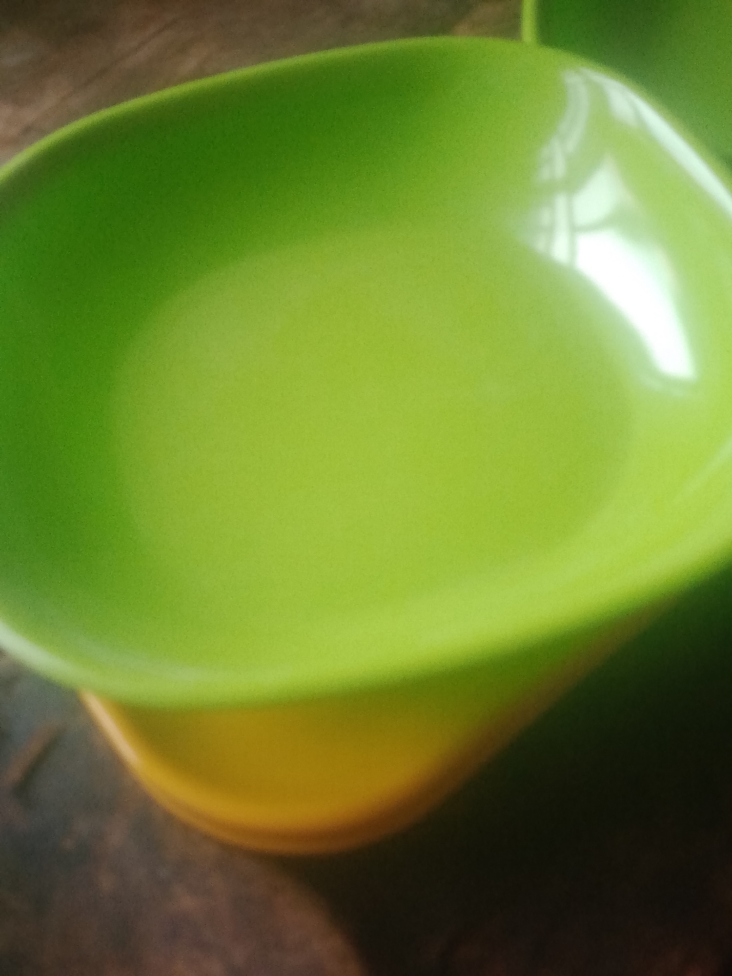 The image shows a close-up, out-of-focus view of a green plastic bowl with a yellow base, resting on a dark, textured surface, possibly wood. The lighting creates bright reflections on the bowl's surface. There are no people or discernible activities depicted in the image. The location in Jauro Yinu, Nigeria, is not evident from the visual information provided.