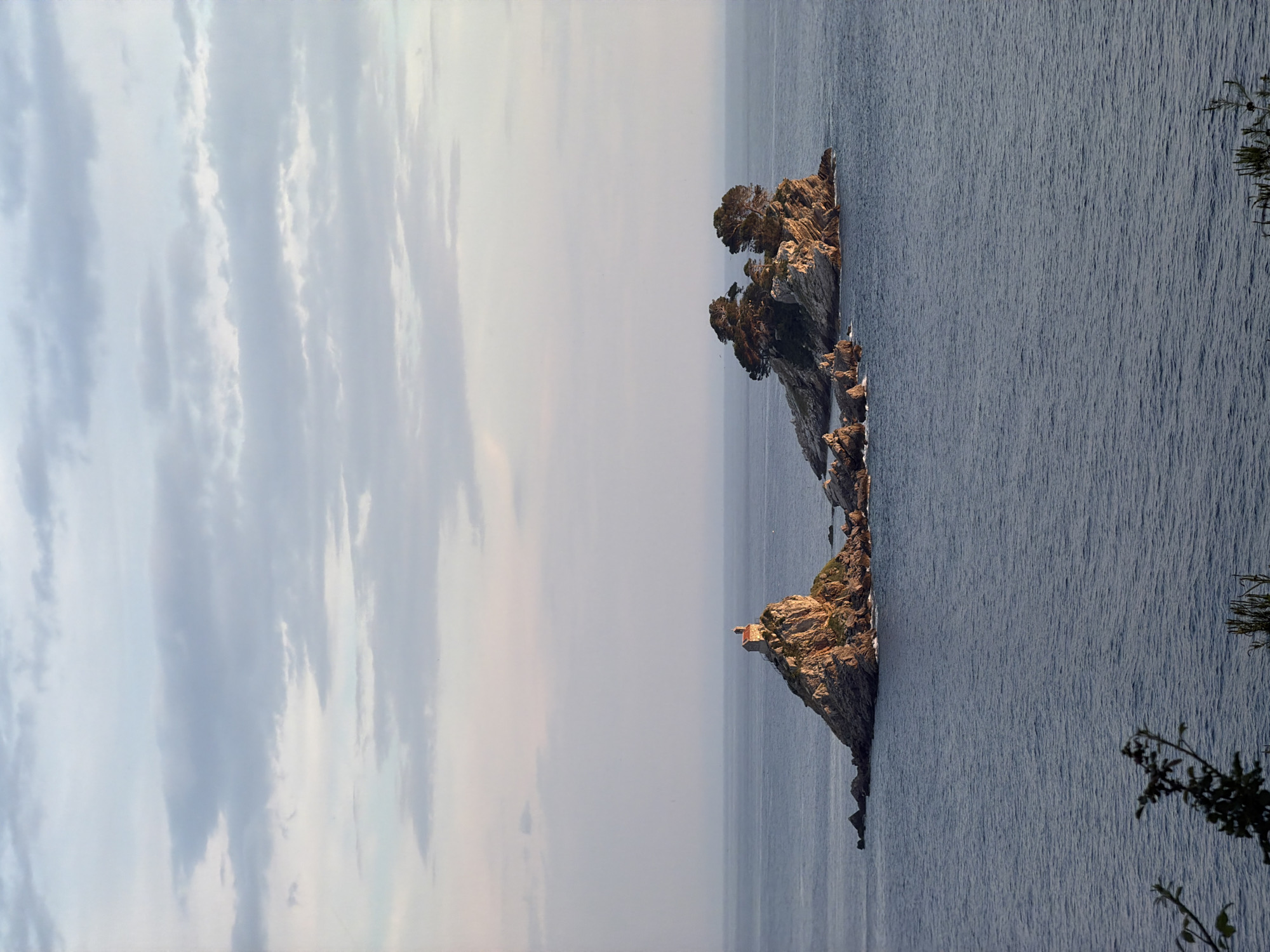 The image depicts two rocky islets in the sea off the coast of Petrovac na Moru, Montenegro. The smaller islet has a small building atop it. The sky is cloudy. The water is visible and calm. Some greenery is visible on the right side.