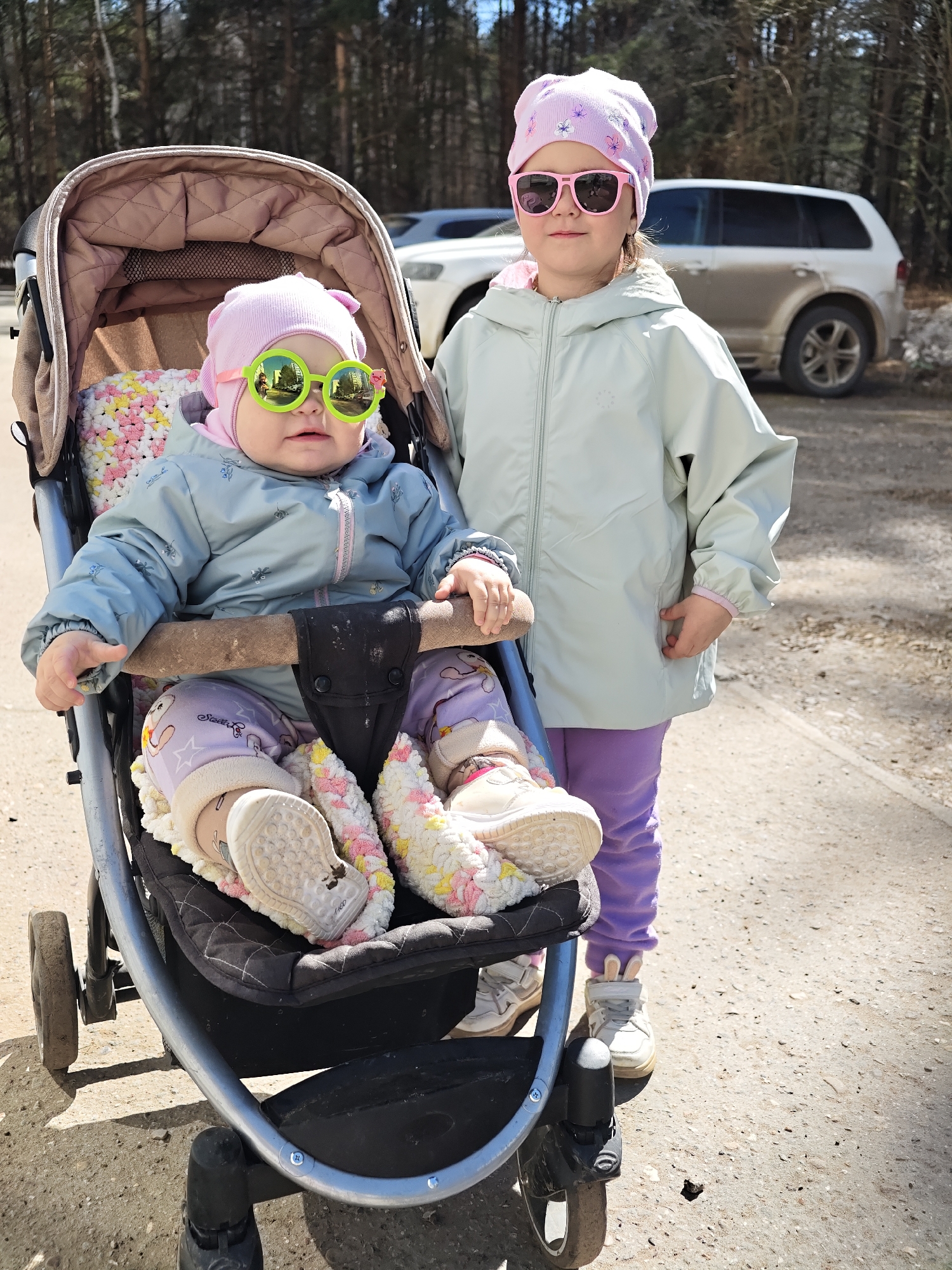 Two young children are outdoors on a partly sunny day in what appears to be a parking area near a forest in Chaikovsky, Russia. A baby, wearing a pink floral beanie, light blue jacket, and patterned leggings with white shoes, sits in a black stroller with a beige canopy. The baby is also wearing bright green sunglasses. A second child, slightly older, stands beside the stroller. This child wears a matching pink floral beanie, pink sunglasses, a light green jacket, and purple leggings with white sneakers featuring rabbit ear details. Two vehicles are parked in the background; a white SUV is partly obscured. The ground is concrete or asphalt with patches of dirt. A forest can be seen in the background.