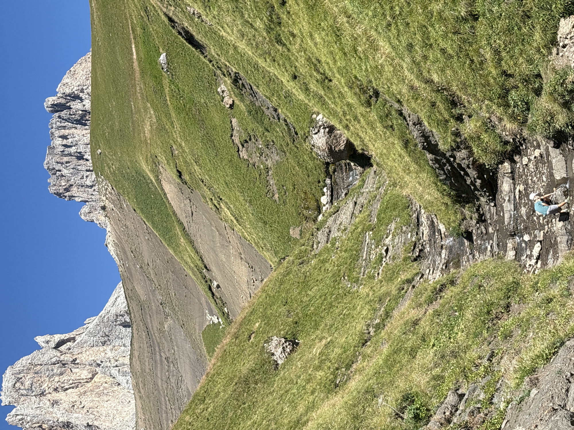 The image depicts a mountainous landscape in Campitello di Fassa, Italy, under a clear blue sky. A narrow stream bed, formed by rocks and water, runs through the center of the frame, descending from the upper part of the image. The surrounding slopes are covered in lush green grass and interspersed with rocky outcrops and patches of dark earth. In the distance, rugged, pale mountains rise prominently against the sky. At the lower part of the stream bed, a person wearing a light-colored hat and a teal shirt is crouched down, appearing to interact with the water. They are accompanied by a black bag. The scene captures a natural, outdoor environment with a single individual engaged in an activity by the stream.