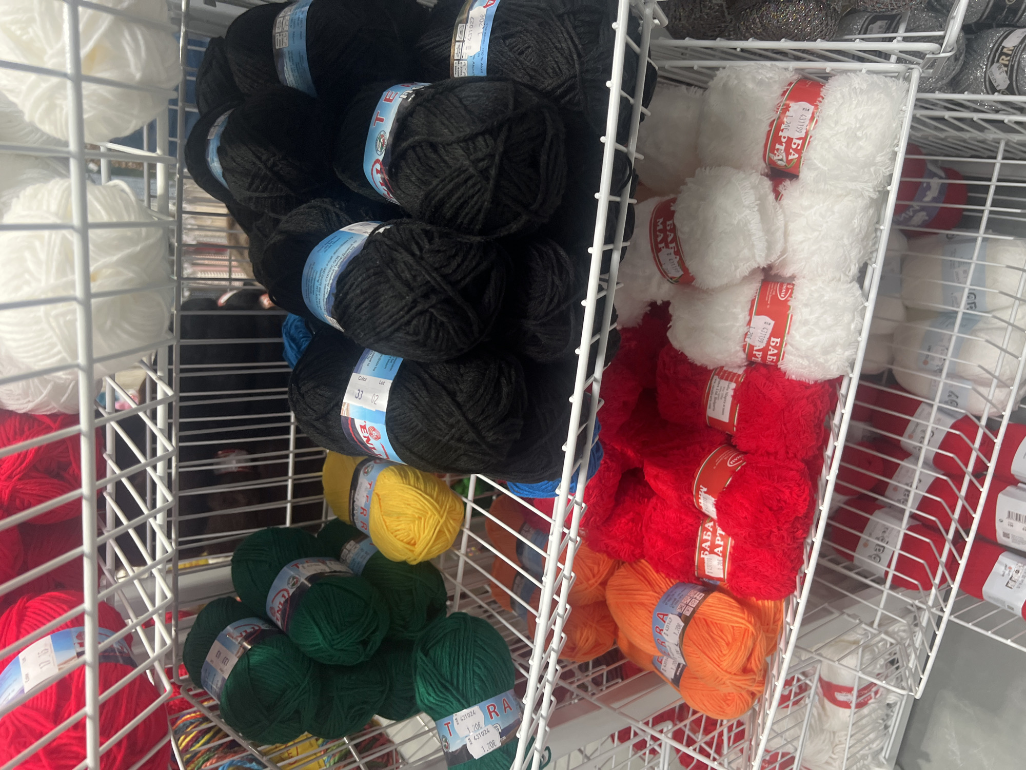 This indoor image shows a retail display of yarn spools in a wire shelving unit. The colors of the yarn include white, black, red, orange, yellow, and green. Several spools have labels with the word "TERRA" visible. The text "Burgas, Bulgaria" is derived from the brand name TERRA, but there is no other information in the image to verify the location. The scene appears to be within a store or shop selling yarn or craft supplies.