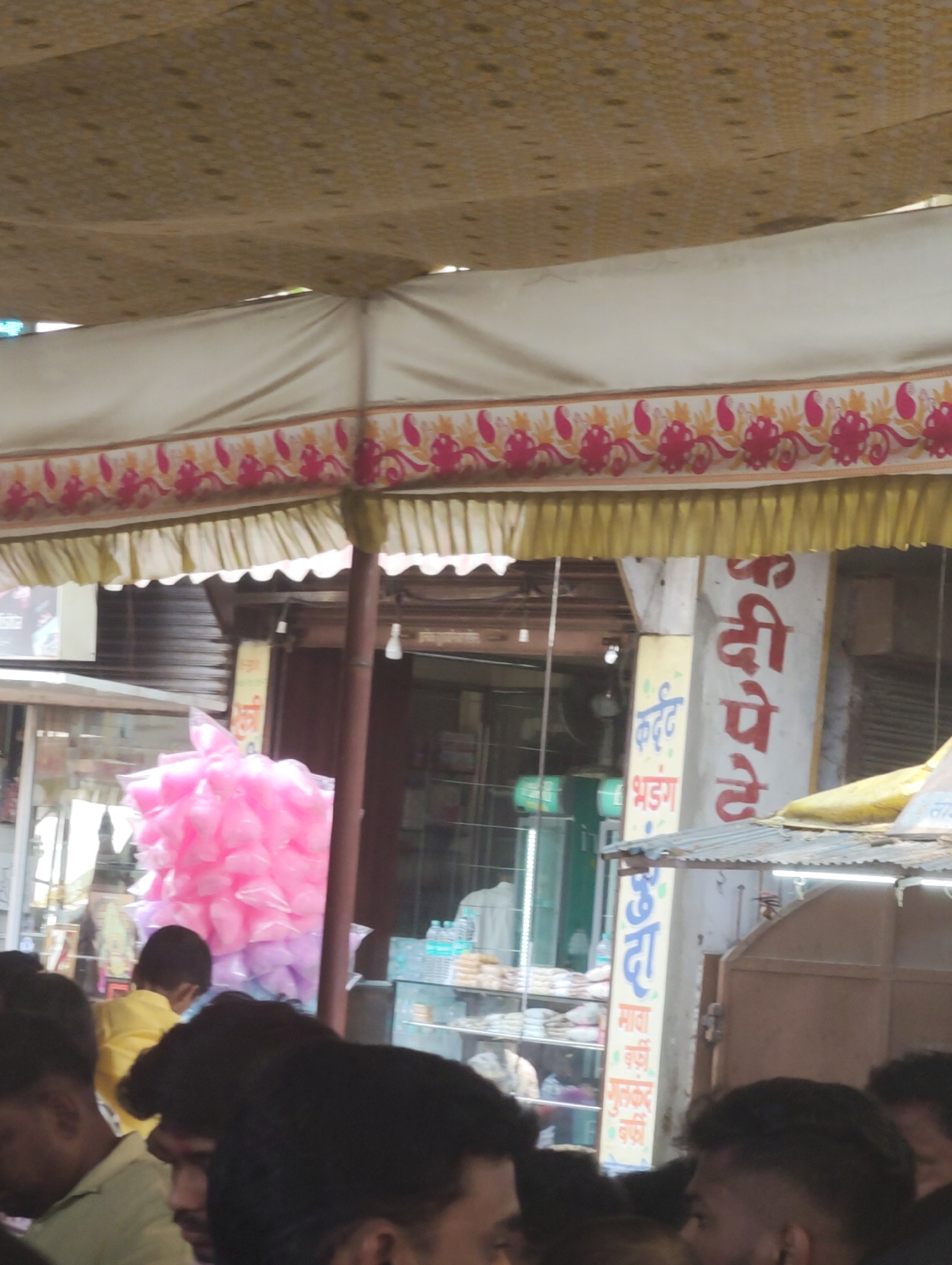 This image depicts a bustling outdoor street scene in a commercial area of Kolhapur, India, likely a market or a street lined with shops. The scene is viewed from under a temporary fabric canopy that covers the upper portion of the frame, featuring a beige patterned top section, a white central strip, and a decorative border with red and yellow paisley/floral motifs, finished with a yellow pleated frill.
In the mid-ground, a prominent display of bright pink cotton candy, individually wrapped in clear plastic bags, is stacked high to the left of the frame, with some purple bags visible at the bottom. Behind this display, a shop interior is visible through an open doorway, showcasing shelves stocked with packaged food items, possibly sweets, and bottled water. A person wearing a white shirt is partially visible inside this shop.
To the right, a white vertical pillar features a sign with large red Marathi text reading "कंदी पेढे" (Kandi Pede), identifying the shop's specialty. Adjacent to this, a narrower yellow sign with blue Marathi text advertises "कर्डंट भडंग" (Kardant Bhadang) and various types of "बर्फी" (Barfi), confirming a sweet shop or snack vendor. Below these signs, a corrugated metal awning provides additional cover. Further to the right, a closed, dark brown wooden door is visible. White light bulbs are suspended from the canopy structure.
The foreground is dominated by a crowd of people, mostly showing the tops of their heads and shoulders, suggesting a busy environment. Their faces are largely obscured or out of focus. One person in the mid-ground, wearing a yellow shirt, is partially visible from behind, looking towards the cotton candy display.