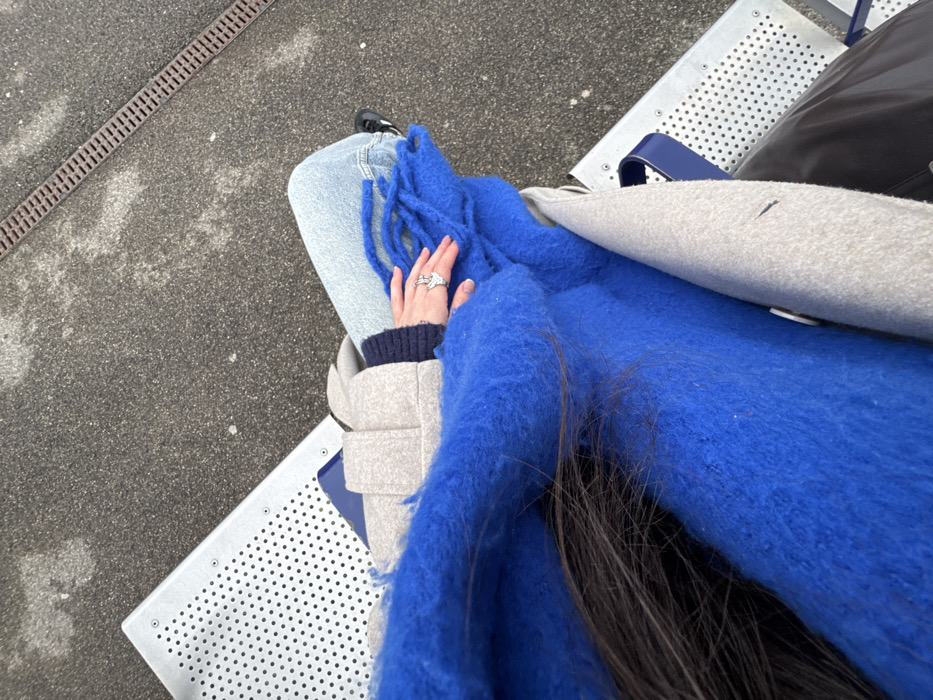 Overhead shot of a person seated outdoors. The person wears blue jeans, a navy sweater, and a light gray coat. A bright blue, fluffy scarf is draped across their lap. The person's left hand, adorned with multiple rings, rests on the scarf. The person sits on a silver, perforated bench with a blue accent. A dark bag sits to the upper right. A concrete surface is visible to the left, with a linear drainage grate. Some strands of long, dark hair are visible beneath the scarf.