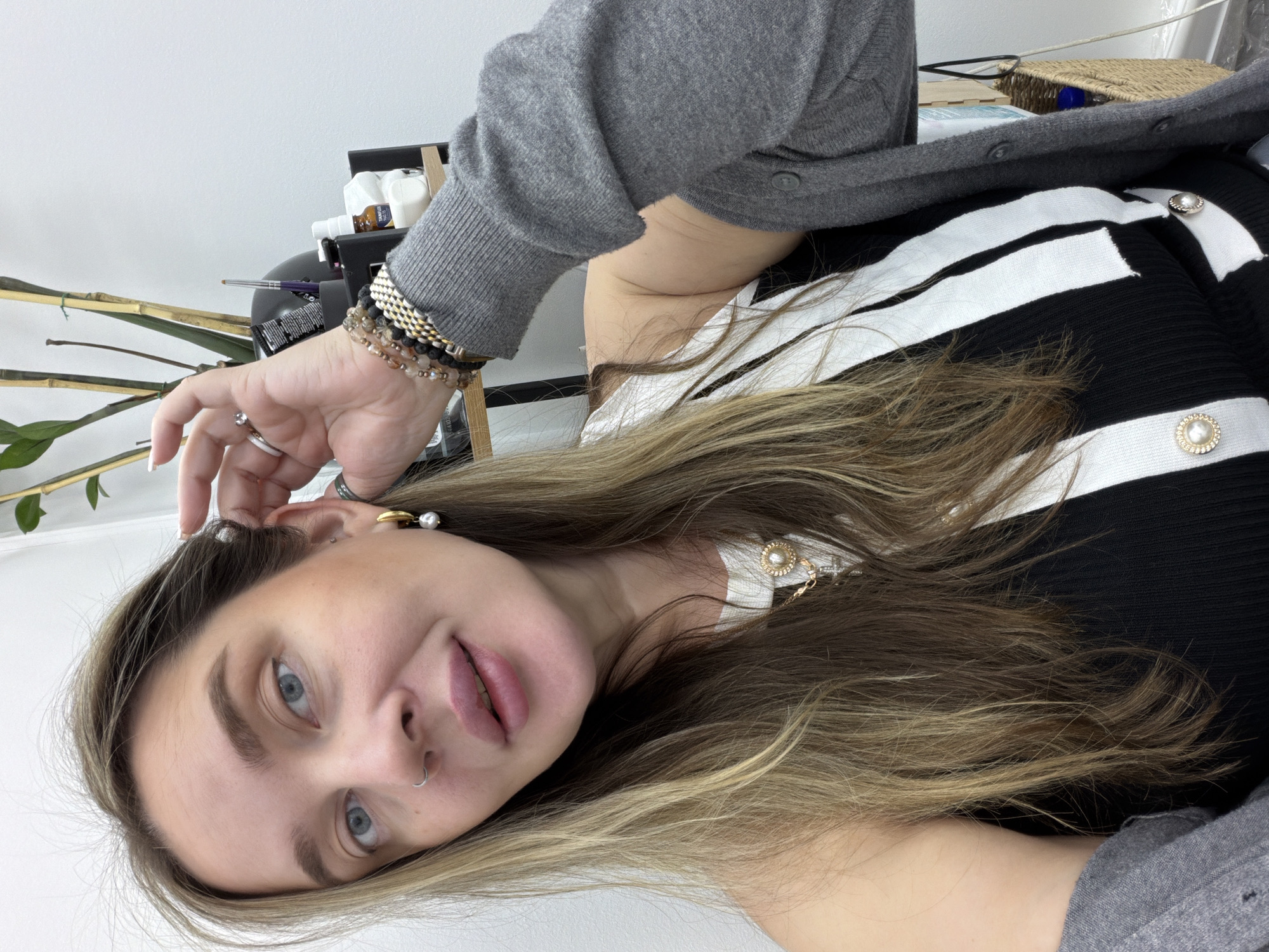 A selfie features a woman with fair skin, light brown hair with blonde highlights, and blue eyes. She is wearing a black and white striped top with pearl-like buttons and a gray cardigan. Multiple bracelets adorn her left wrist, and she has a silver ring on her finger. She is touching her left ear, which is adorned with earrings. The background is a white wall with a small plant and some items on a shelf. The location in Moscow, Russia is not discernible from the image.