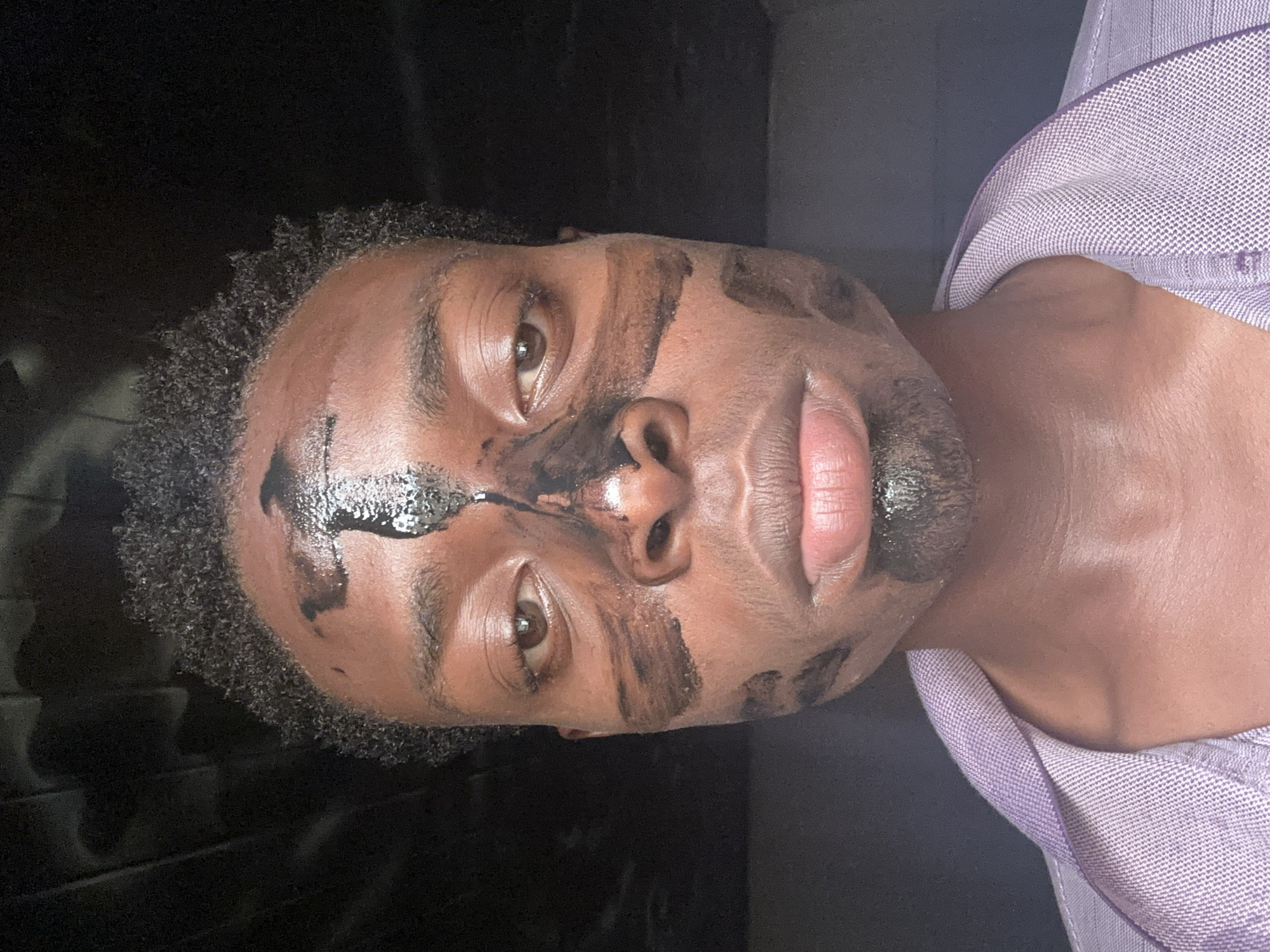 A man with dark curly hair is lying on his back, facing the camera. He is wearing a light purple shirt. His face is covered with a black substance resembling a facial mask, applied unevenly across his forehead, nose, cheeks, and chin. The background is dark and indistinct. There is no information to indicate the location is Bukuru, Nigeria.