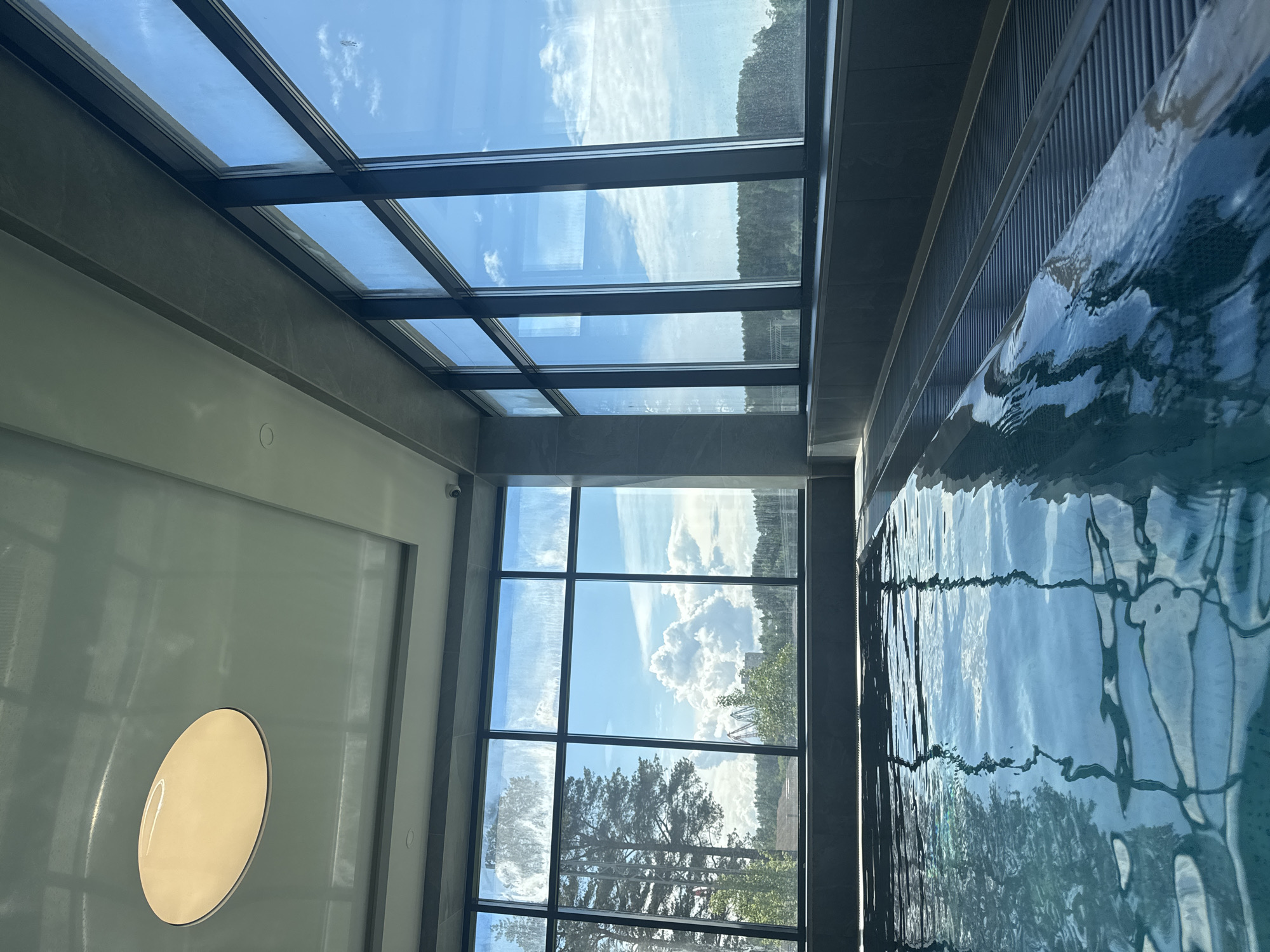The image depicts an indoor swimming pool. The pool's water reflects the sky and trees visible through a large window and a skylight. The window is a grid of glass panes. A circular light fixture is visible on the wall. The location is Chaikovsky, Russia.